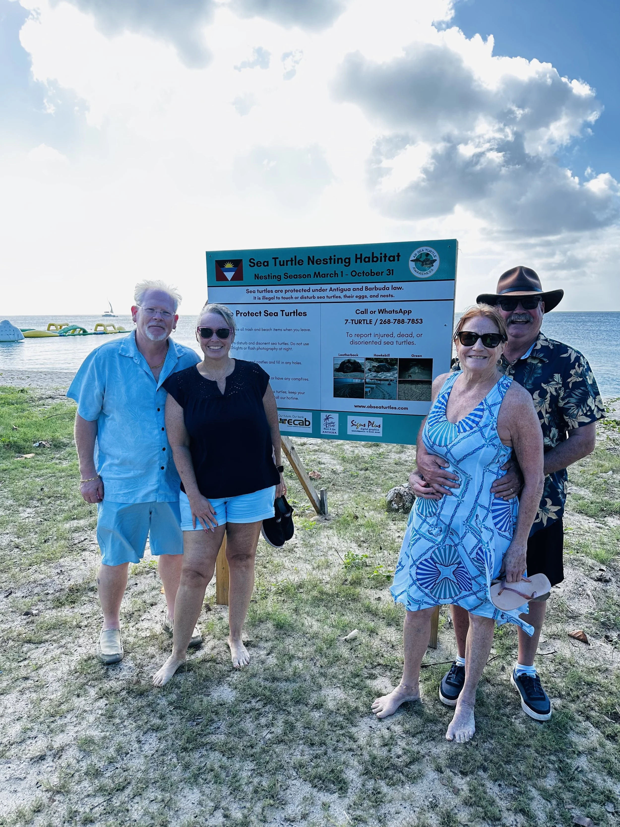 Four people standing in front of a sign about sea turtle nesting habitat on a beach with water and sailboats in the background.