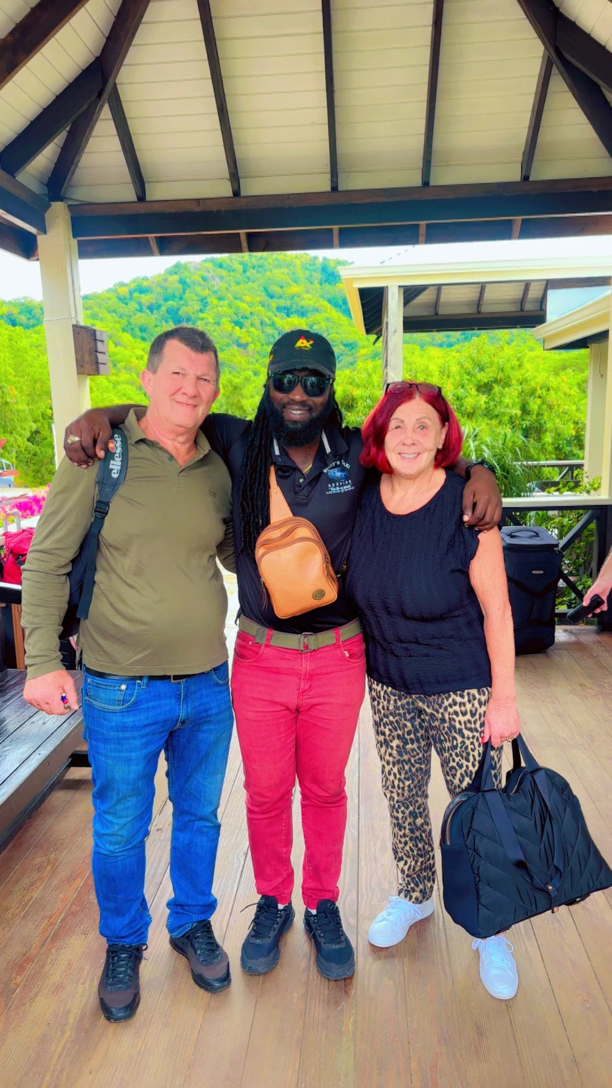 Three people standing together on a wooden deck, smiling, with a backdrop of lush green hills and trees. The person on the left is wearing a light brown long-sleeve shirt, blue jeans, and carrying a backpack. The person in the middle has long dreadlo