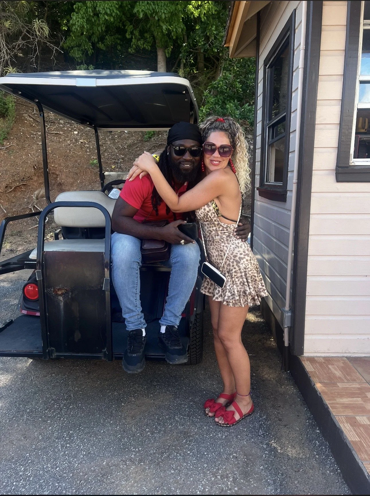 A man and a woman hugging and smiling next to a golf cart, standing outside a building with trees in the background.