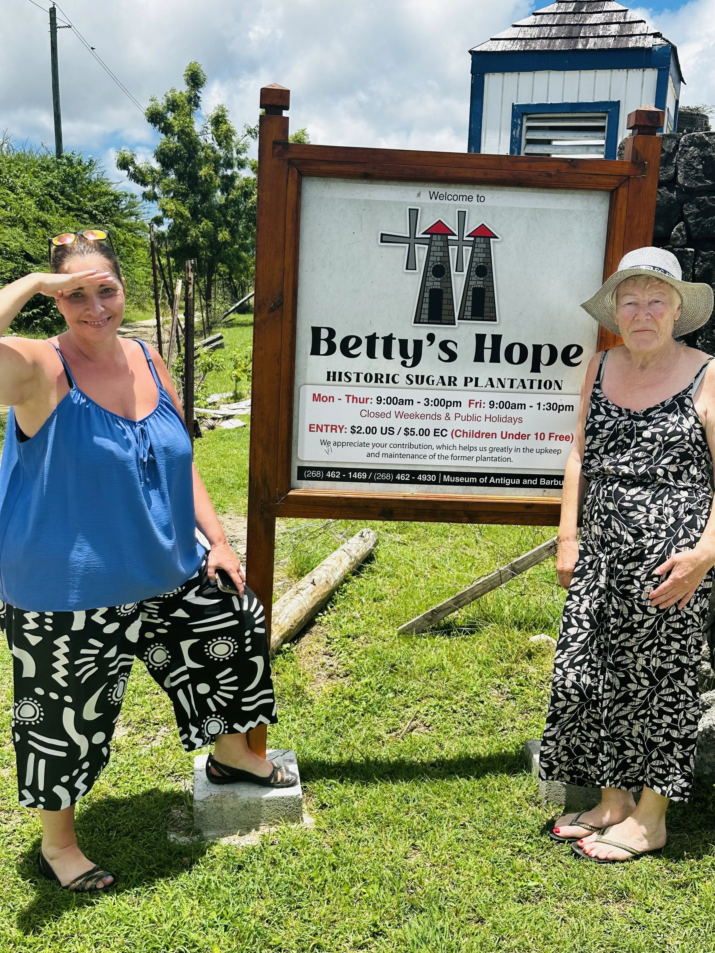 Two women standing next to a sign at Betty's Hope historic sugar plantation in Antigua. One is saluting shade on her eyes, and the other is wearing a wide-brimmed hat.