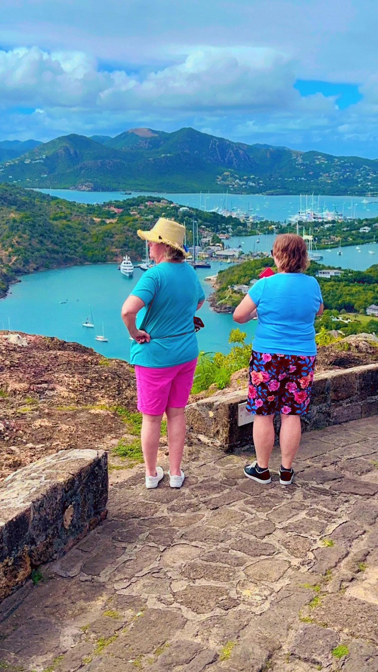 Two women standing on a stone viewpoint overlooking a scenic harbor with boats, mountains, and a partly cloudy sky.