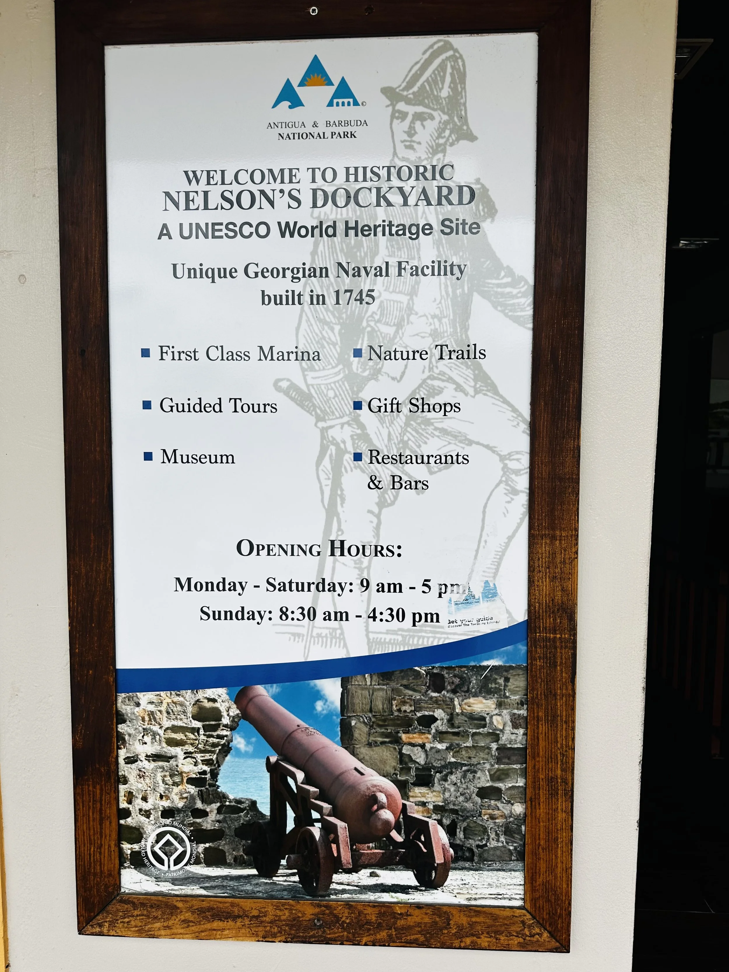 Signboard at Nelson's Dockyard in Antigua and Barbuda National Park with historical information about the site, opening hours, and a picture of an old cannon.