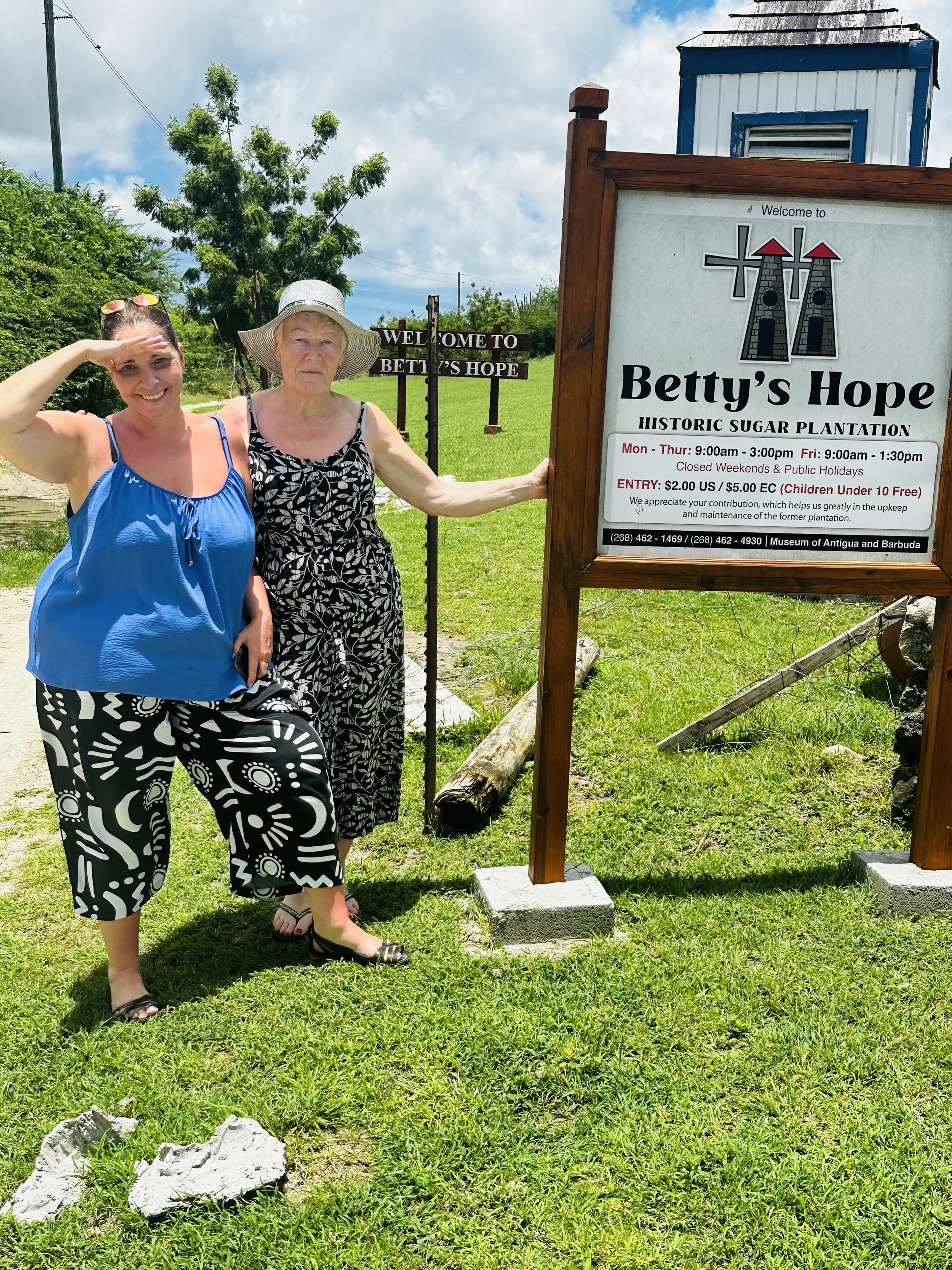 Two women standing outdoors near a sign that reads 'Betty's Hope, Historic Sugar Plantation.' The woman on the left is saluting and wearing a bright blue top and patterned black-and-white pants. The woman on the right is wearing a sun hat and a simil