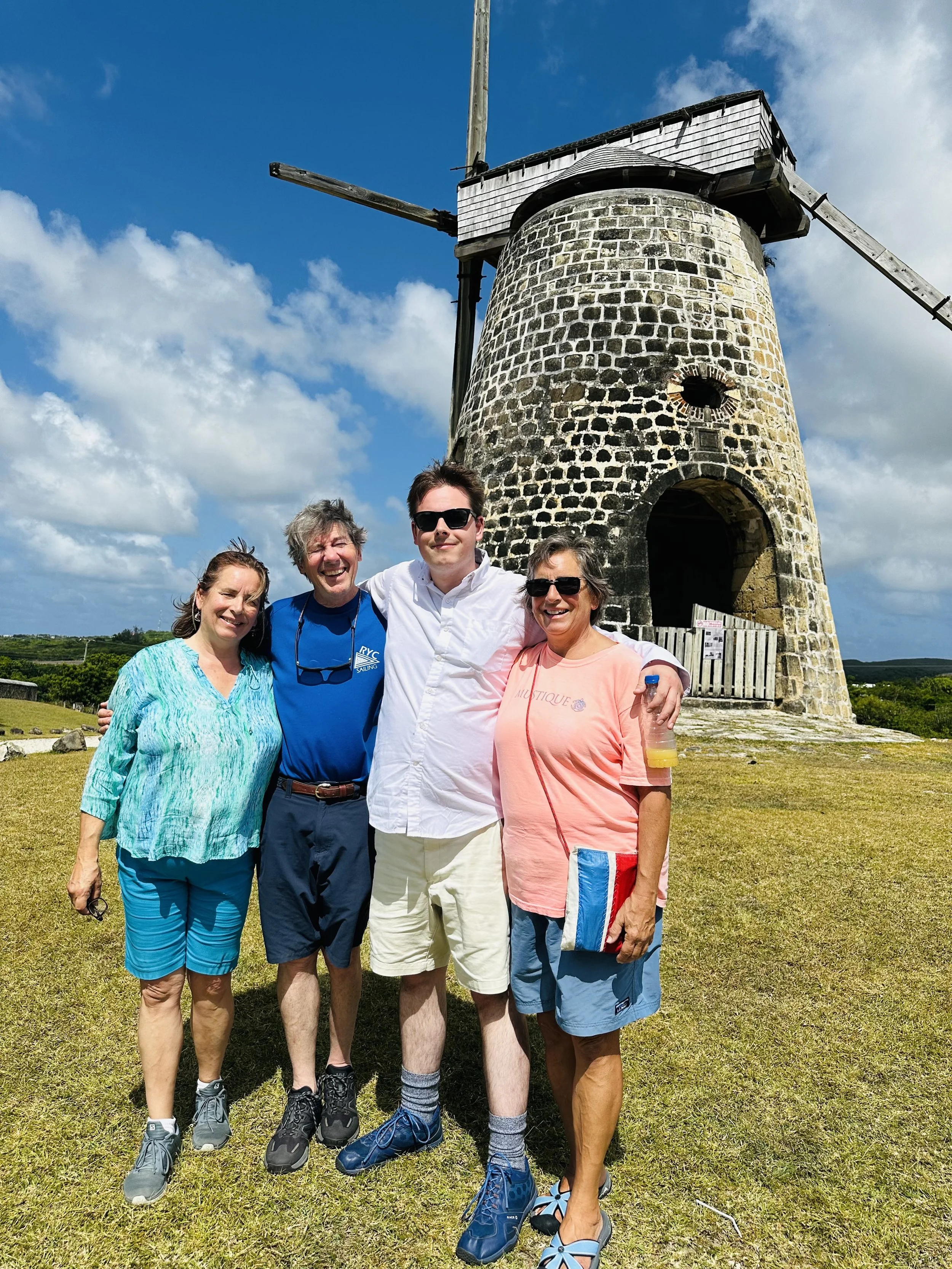 Four people smiling and standing together outdoors in front of a small old stone windmill, on a grassy field with blue sky and clouds overhead.