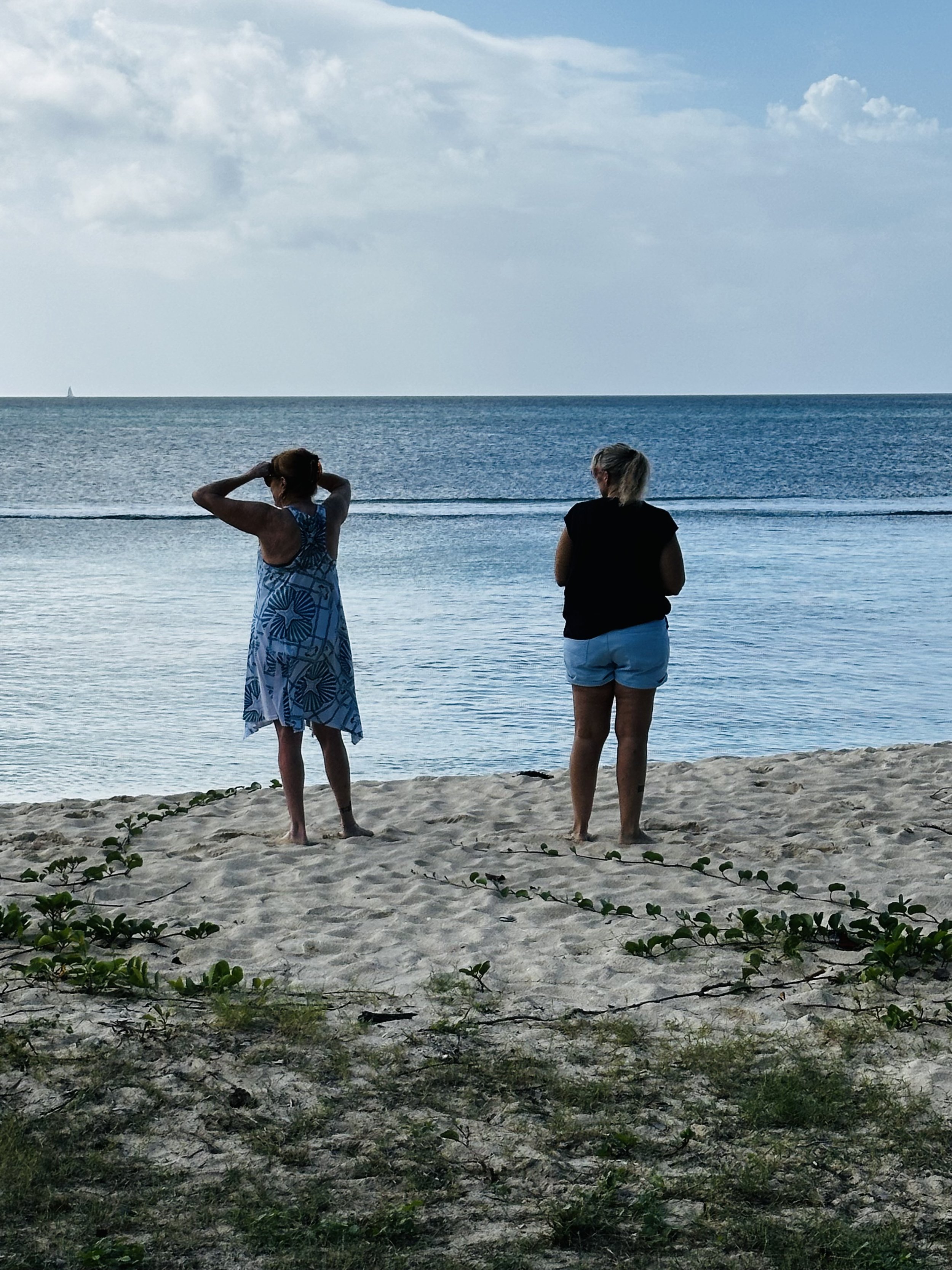 Two women standing on the sandy beach facing the ocean, one in a blue dress and the other in a black top and shorts, with a sailboat in the distance under a partly cloudy sky.