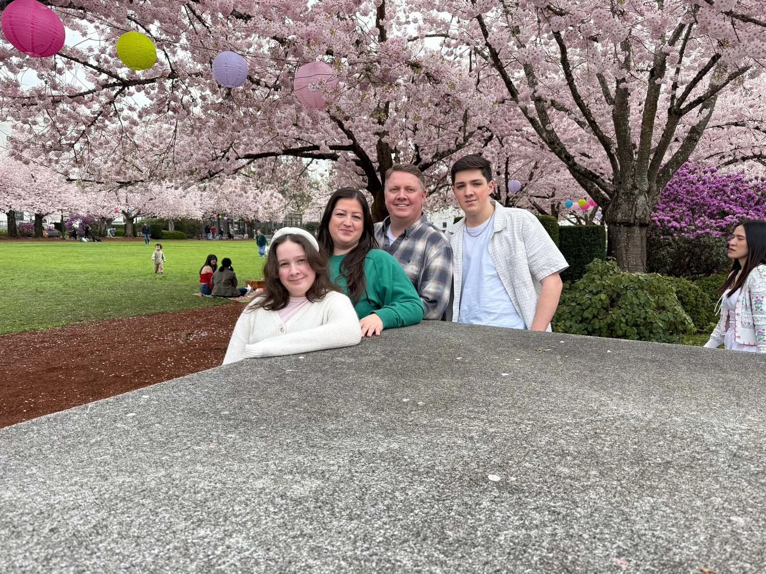 Michael Welsh and his Family of four posing in front of blooming cherry blossom trees with pink flowers, in a park during daytime.