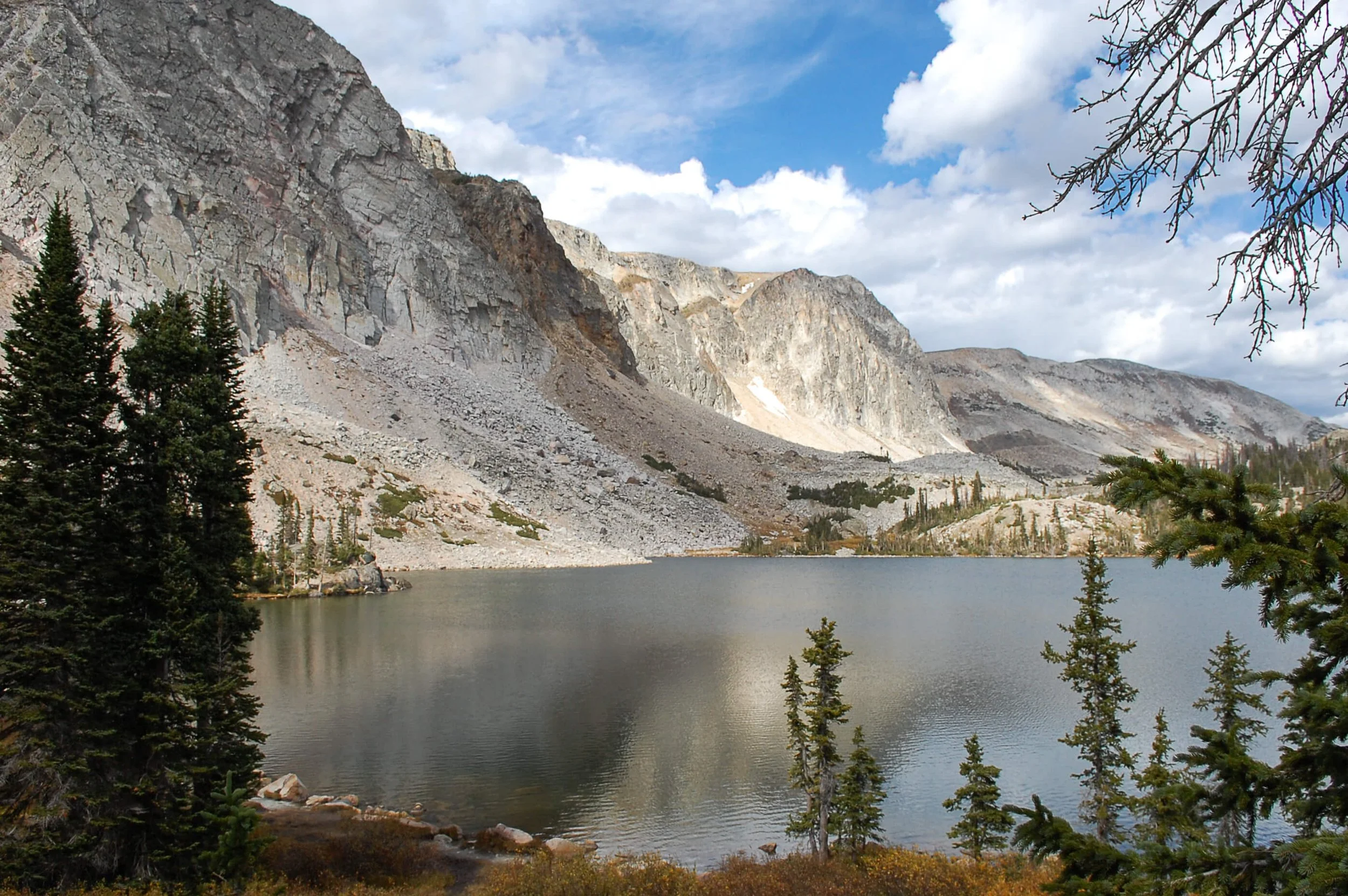 A mountain lake surrounded by pine trees with rocky, gray mountain cliffs in the background under a partly cloudy sky.