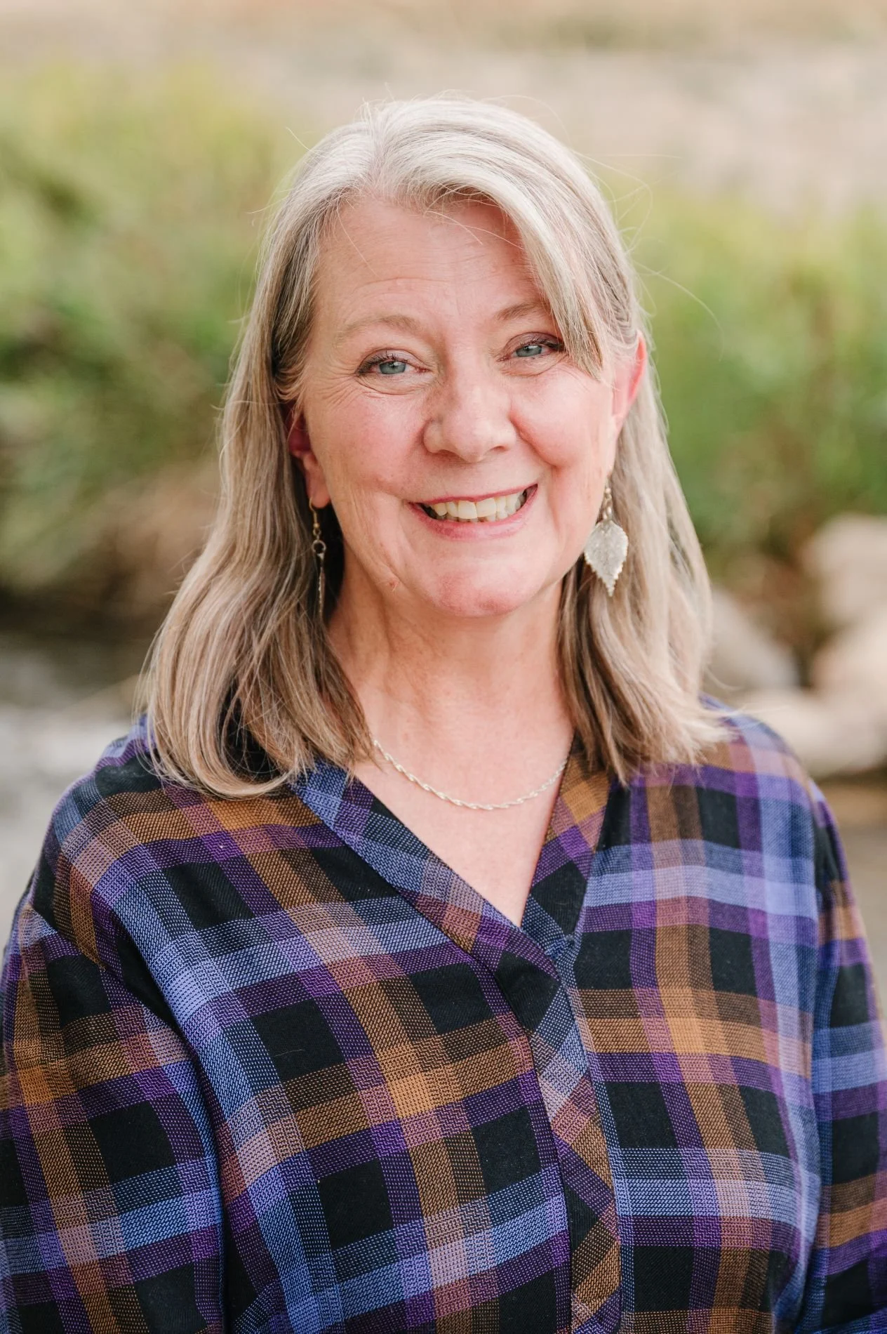 A smiling woman with shoulder-length blonde hair, wearing earrings, a necklace, and a plaid shirt in shades of purple, brown, and black, standing outdoors with blurred greenery in the background.