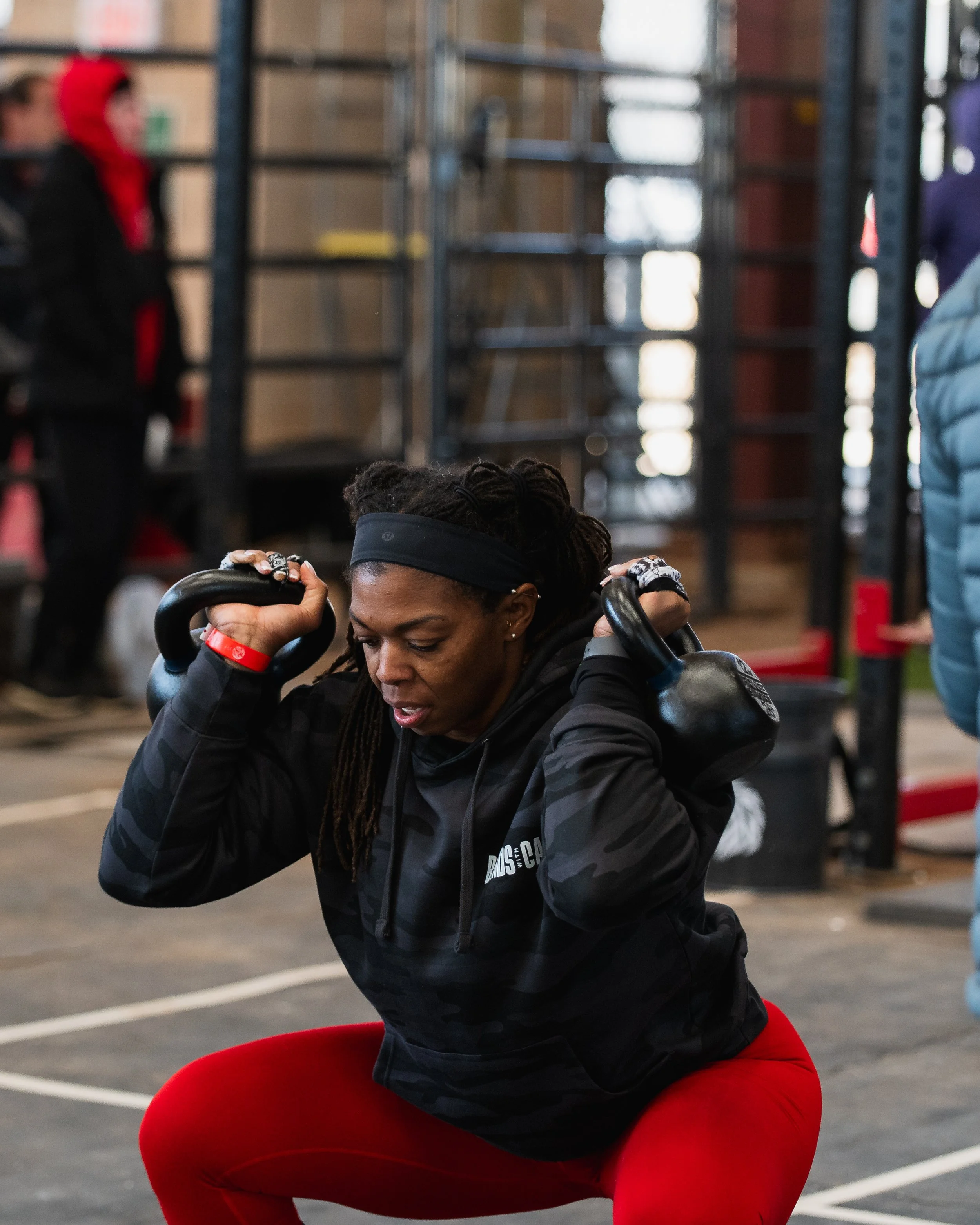 Woman exercising with kettlebell in gym, squatting with bent knees, wearing black jacket with logo, red leggings, and headband, with industrial gym background.