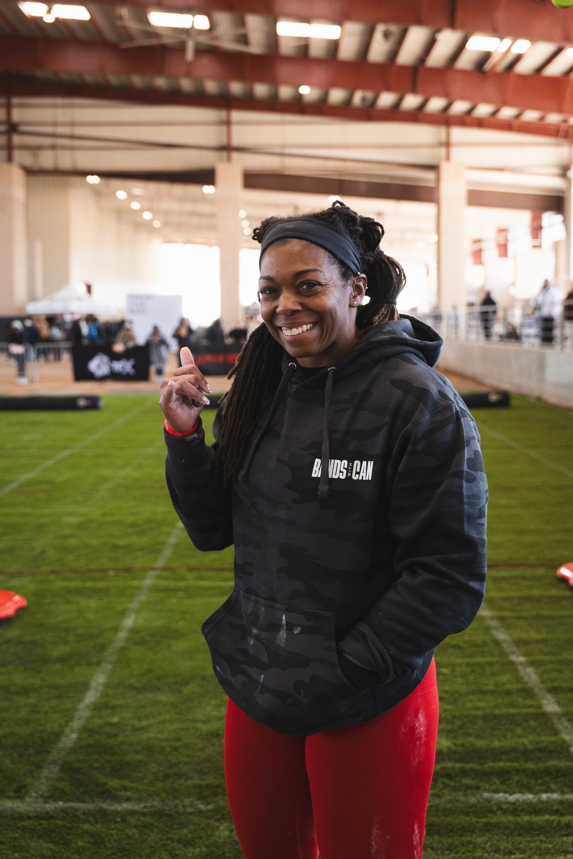 Smiling woman with dreadlocks wearing a black headband and black camouflage hoodie, pointing with her right hand, standing inside a sports facility with artificial turf and blurred background of people and banners.