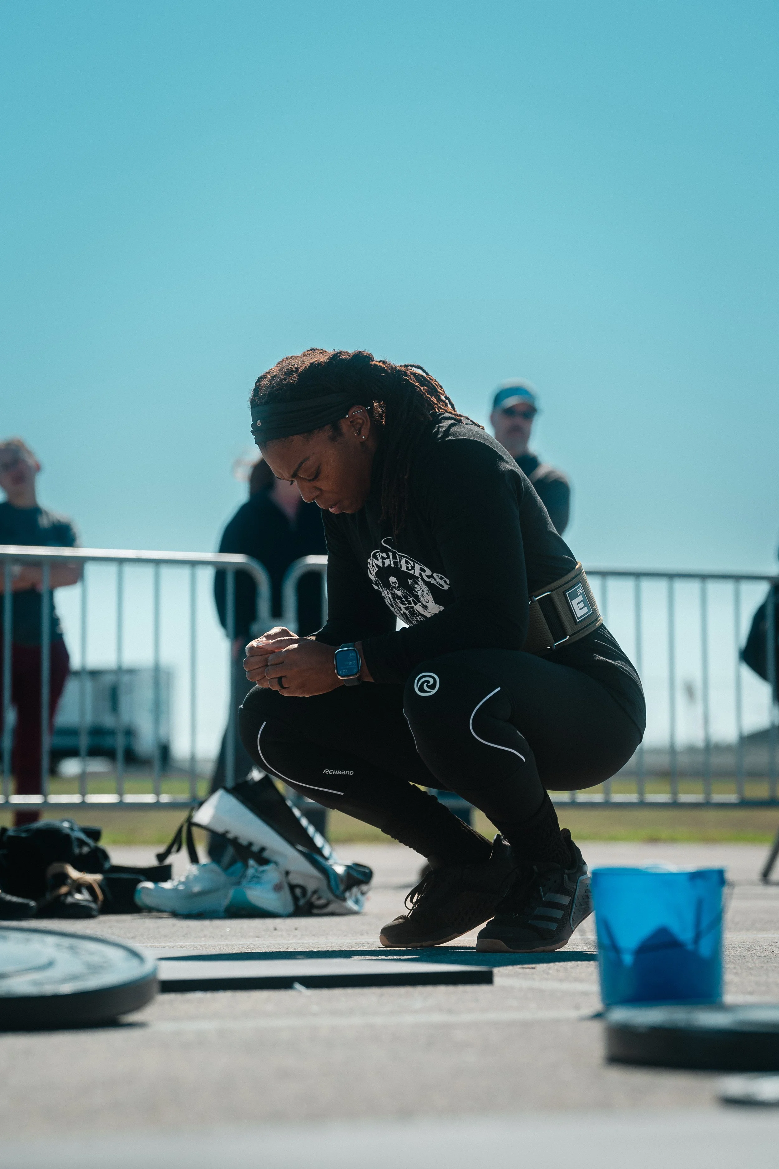 Female athlete crouching on ground, head bowed, wearing athletic clothes and a smartwatch, with people and barriers in the background under a clear blue sky.
