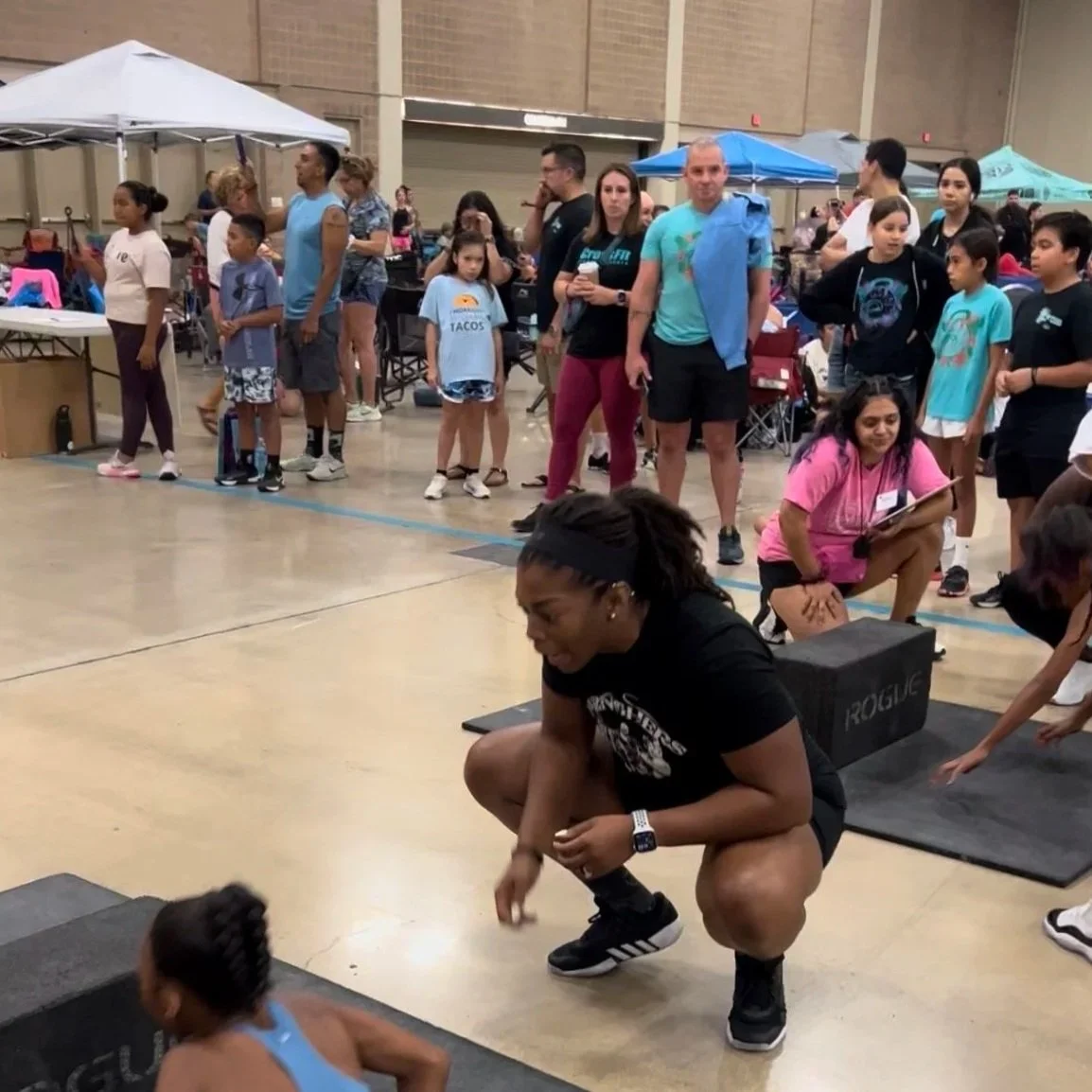 A woman crouching down and encouraging a young girl during a fitness or CrossFit competition in an indoor gym with several spectators and booths in the background.