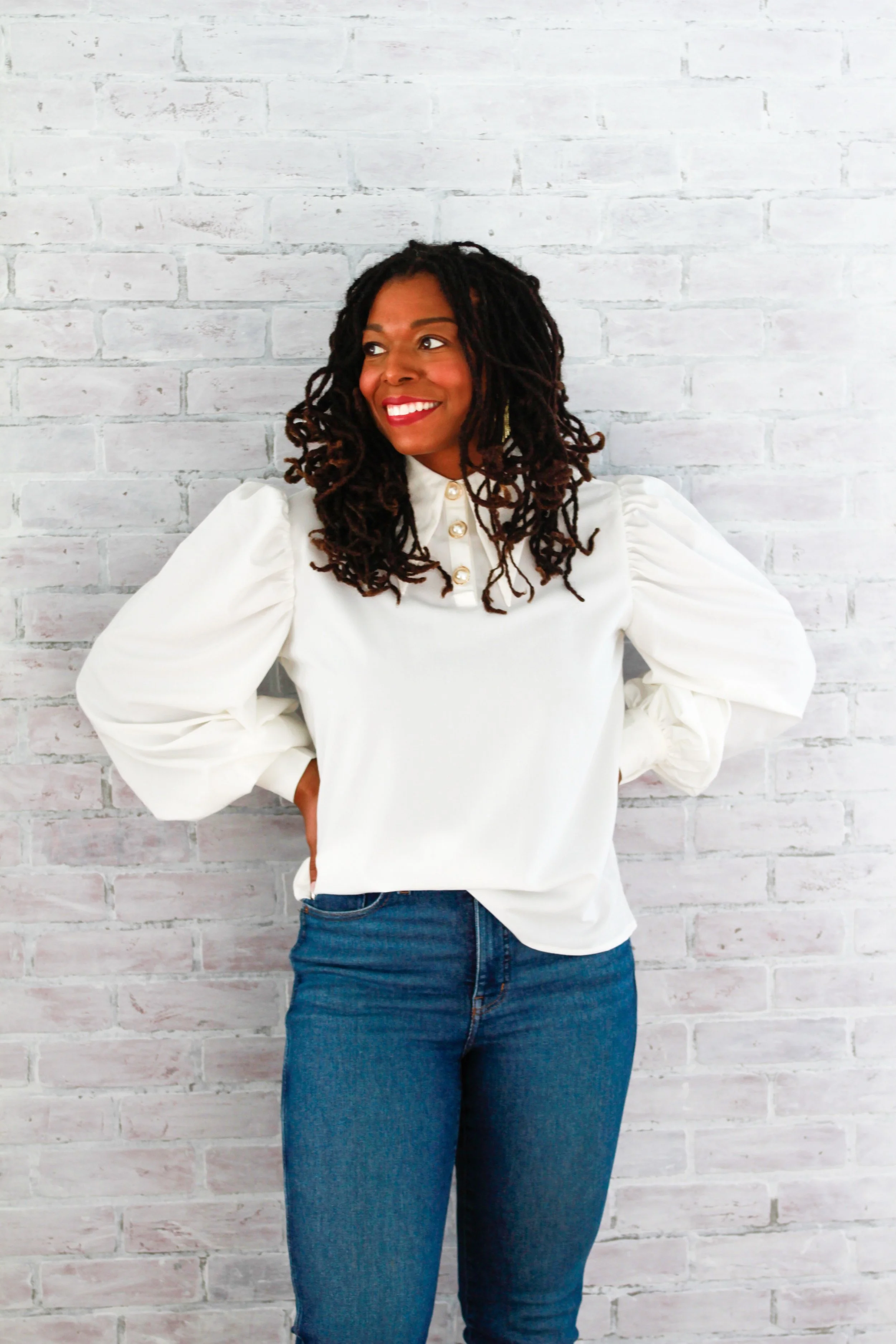 A woman standing against a white brick wall, smiling and looking to her right, wearing a white blouse with puffed sleeves and gold buttons, and blue jeans.