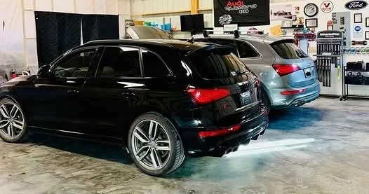 Two parked cars, a black sedan and a gray SUV, inside a garage or service area with automotive tools and equipment in the background.