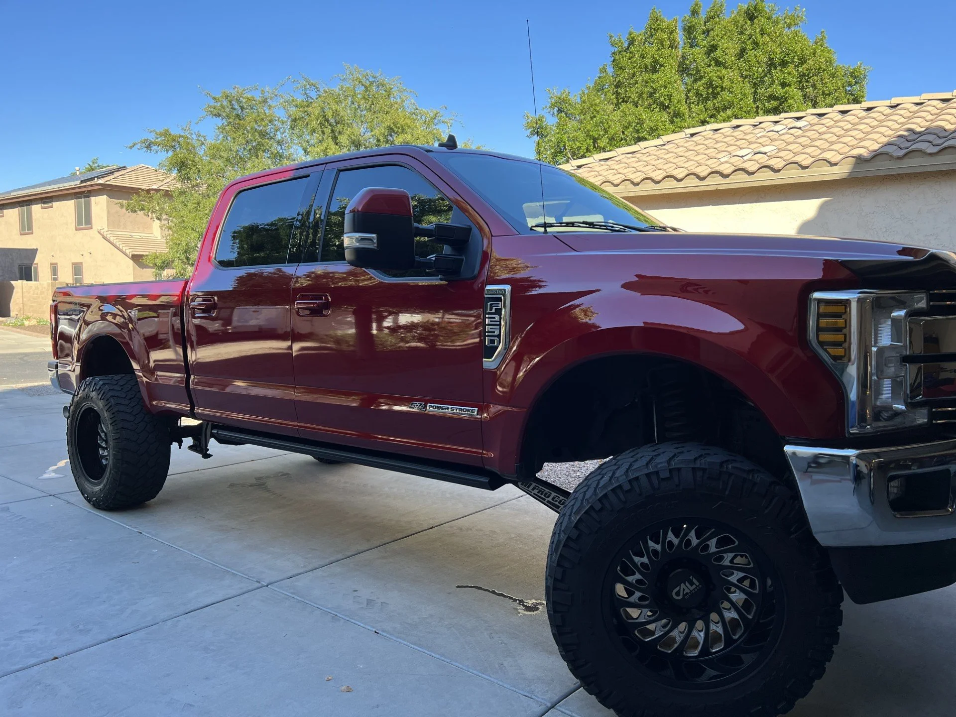 A red Ford F-250 pickup truck with black wheels and off-road tires parked on a driveway in a suburban neighborhood with houses and trees in the background.