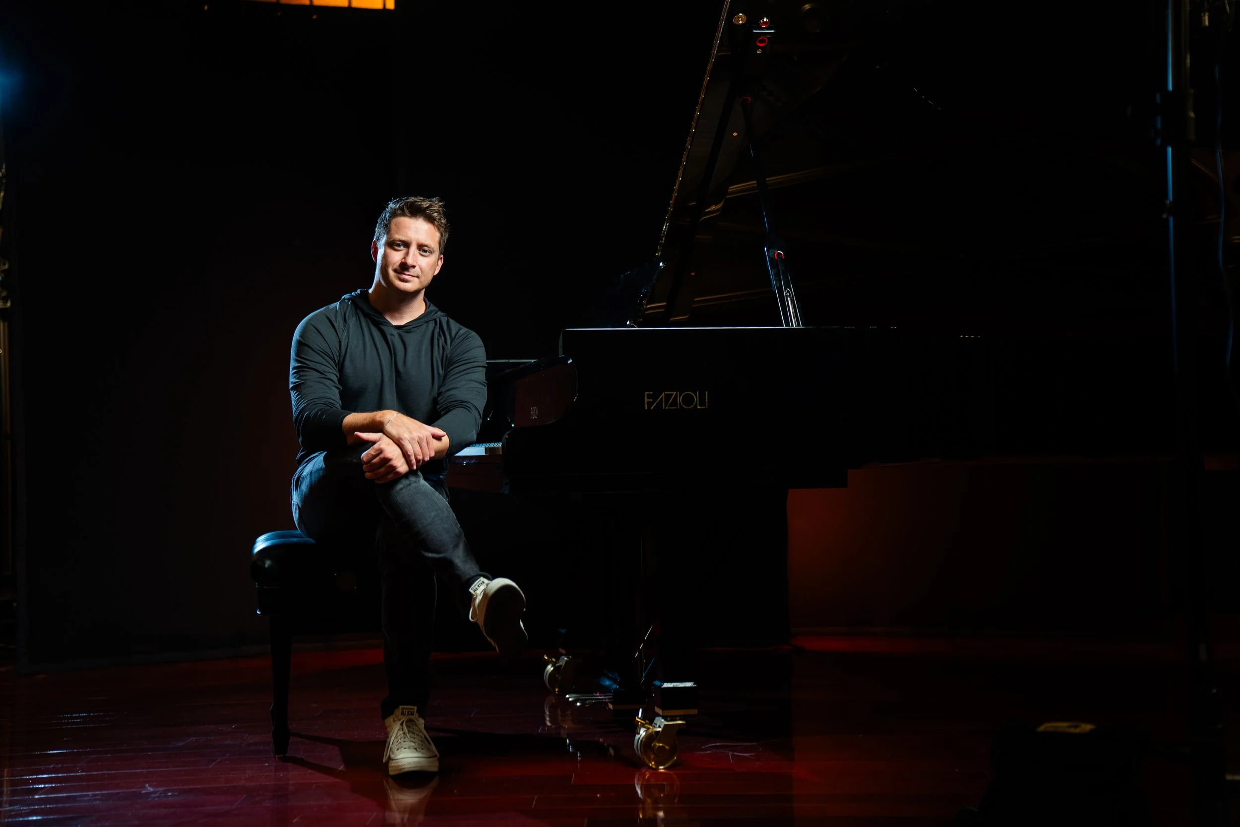 A young man sitting on a piano bench next to a grand piano on a dark stage, illuminated by a spotlight.