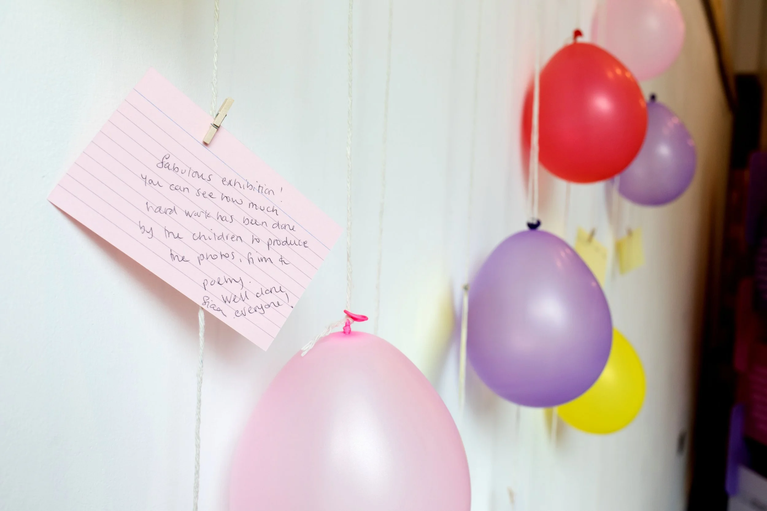 Colorful balloons hanging on a string with pink, purple, yellow, and red colors and a handwritten note pinned to the wall.