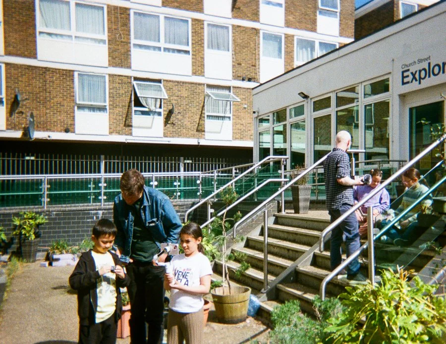 Children and adults gathered outside a building with large windows, some seated on steps and others standing nearby, engaging in activities.