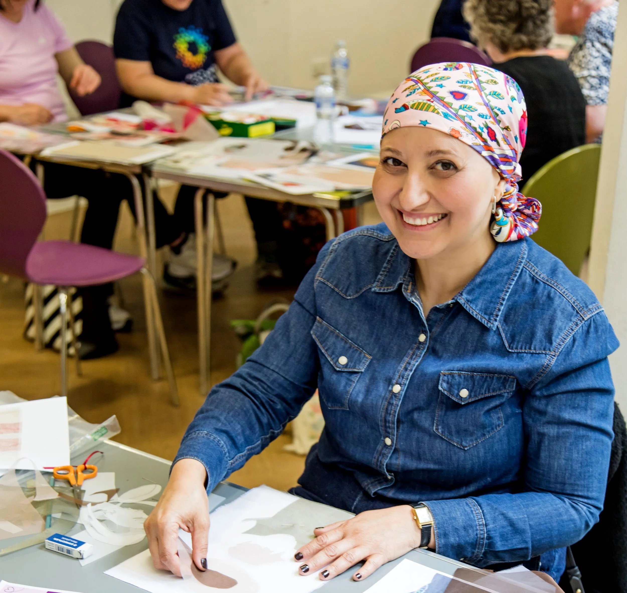 Woman wearing a colorful headscarf and denim shirt smiling while working on an arts and crafts project at a table in a group setting.