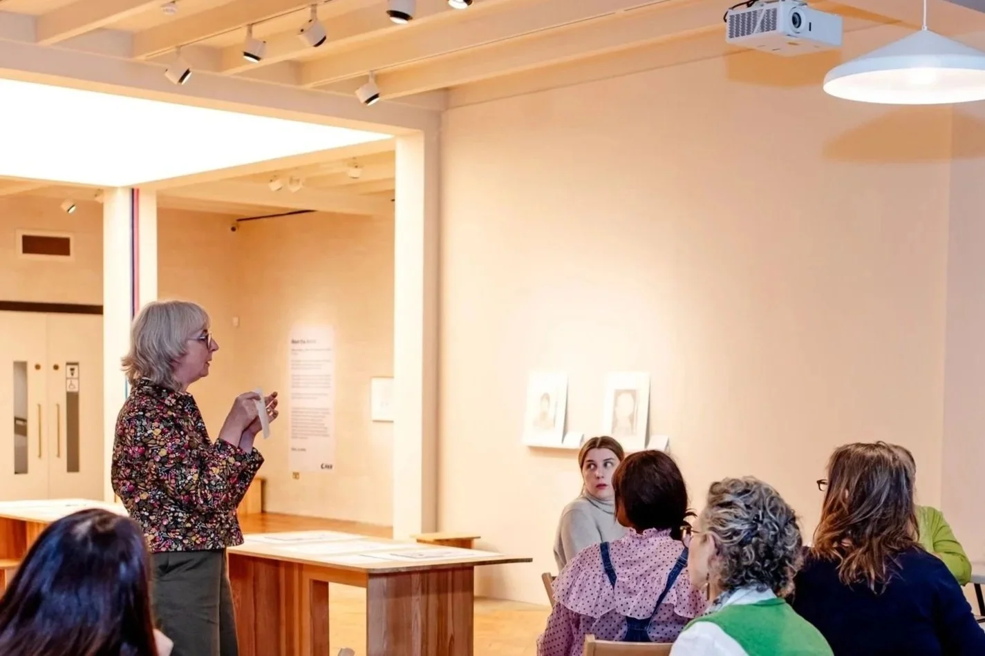 A woman giving a presentation or talk to a small audience in a gallery or classroom setting.