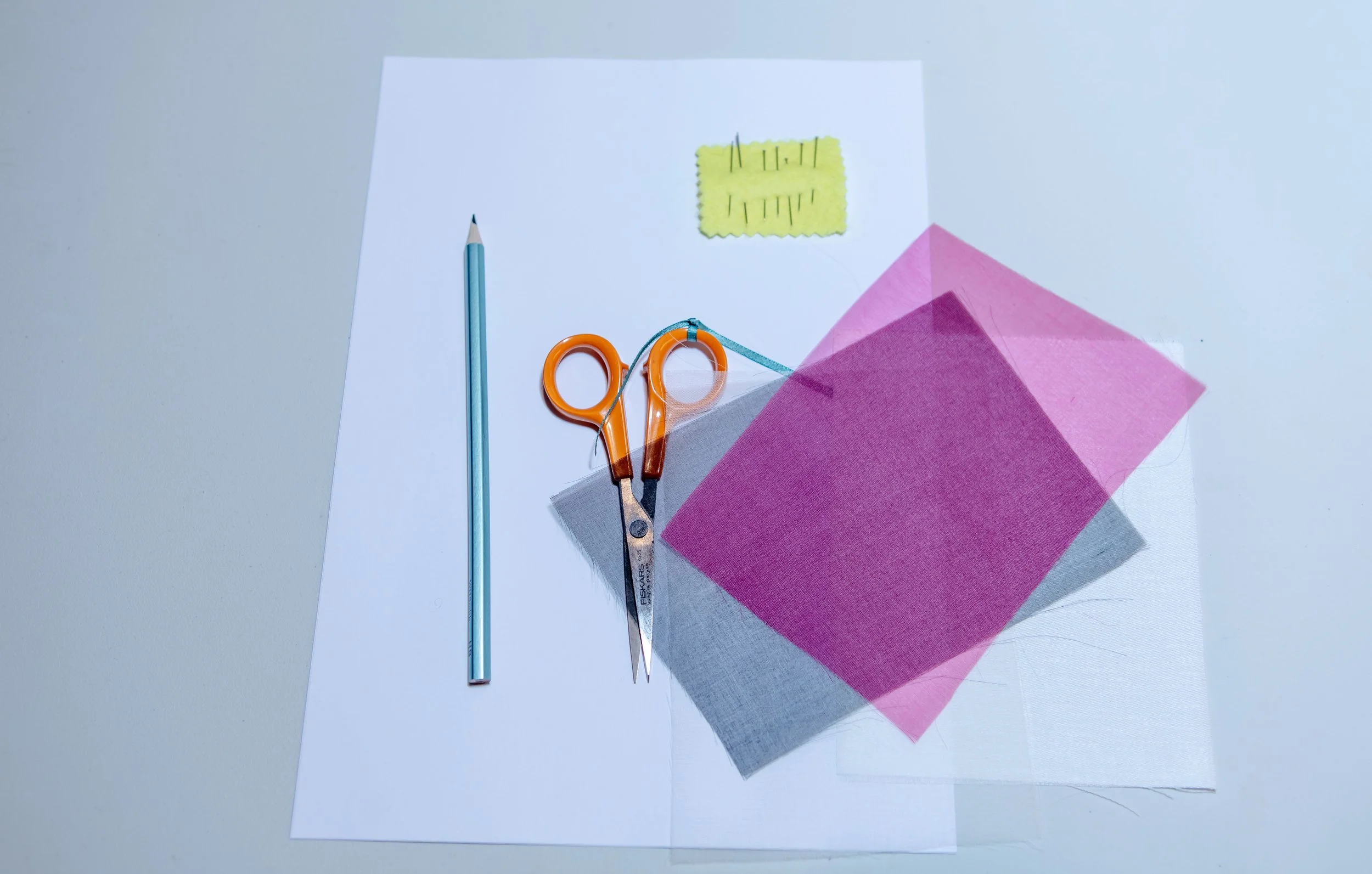 A pair of orange scissors, a blue pencil, various pieces of pink, gray, and white fabric, a piece of yellow felt, and a piece of white paper with a yellow patch, all arranged on a white surface.
