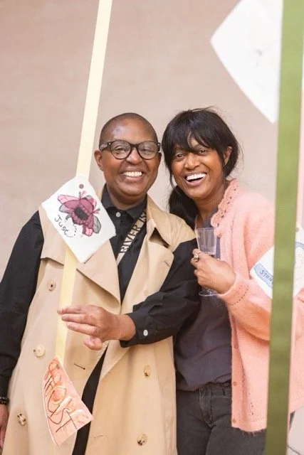 Two women smiling and standing close together at an event, holding a sign with a ladybug illustration and the word 'Usee!' written on it.