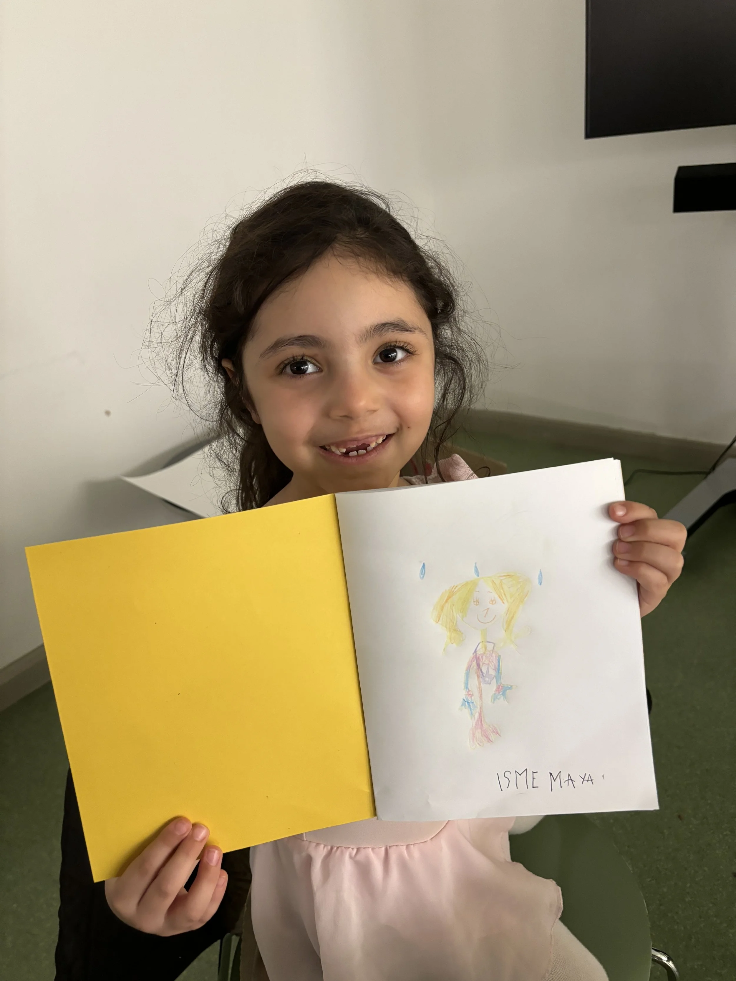 A young girl with dark hair smiling and holding up a drawing of a girl, labeled 'ISME MAX,' in a room with green flooring and white walls.
