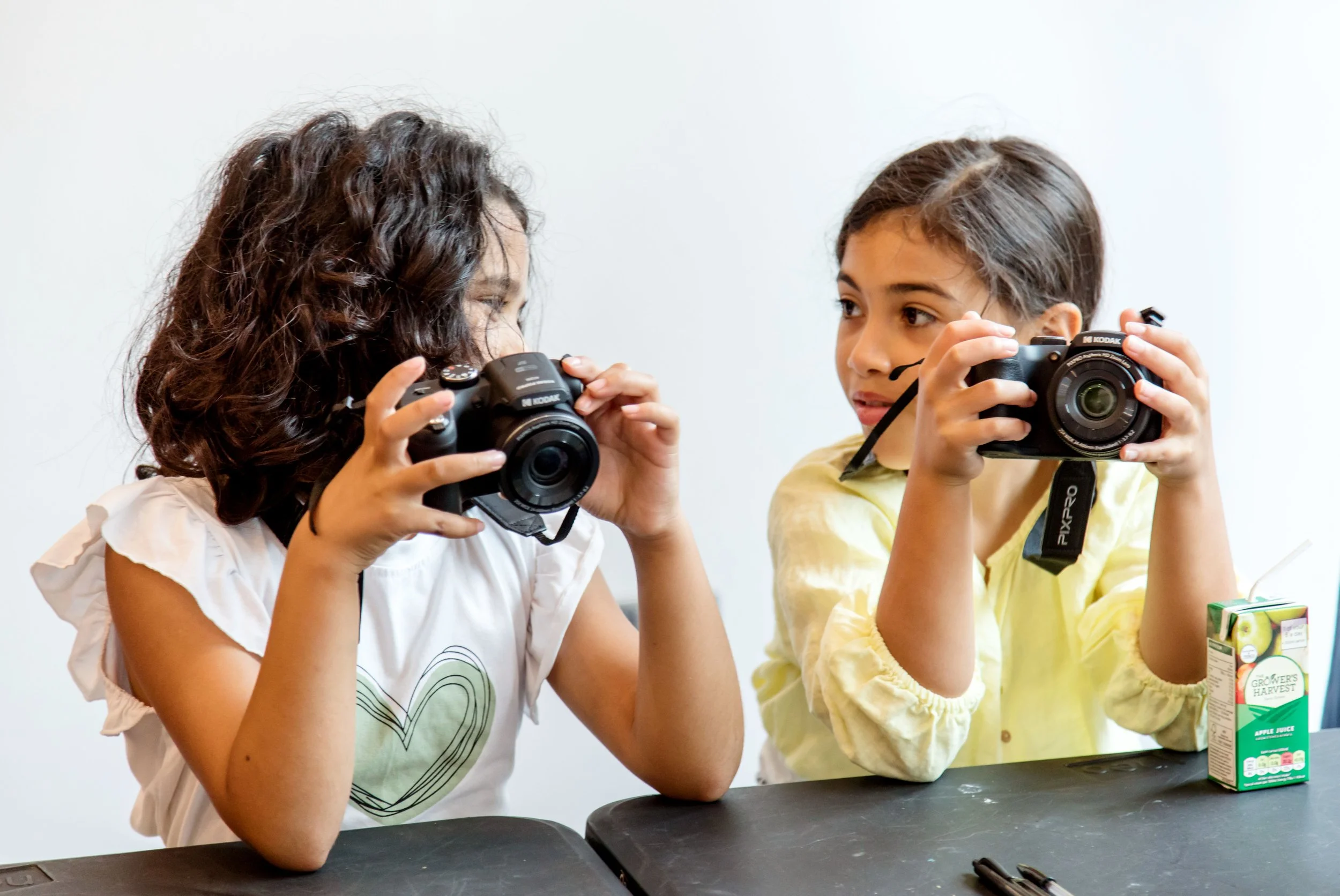 Two young girls sitting at a table, each holding a camera, engaging in photography. One girl has curly hair and wears a white top with a heart design, the other has straight hair and wears a yellow shirt. A carton of apple juice is on the table.