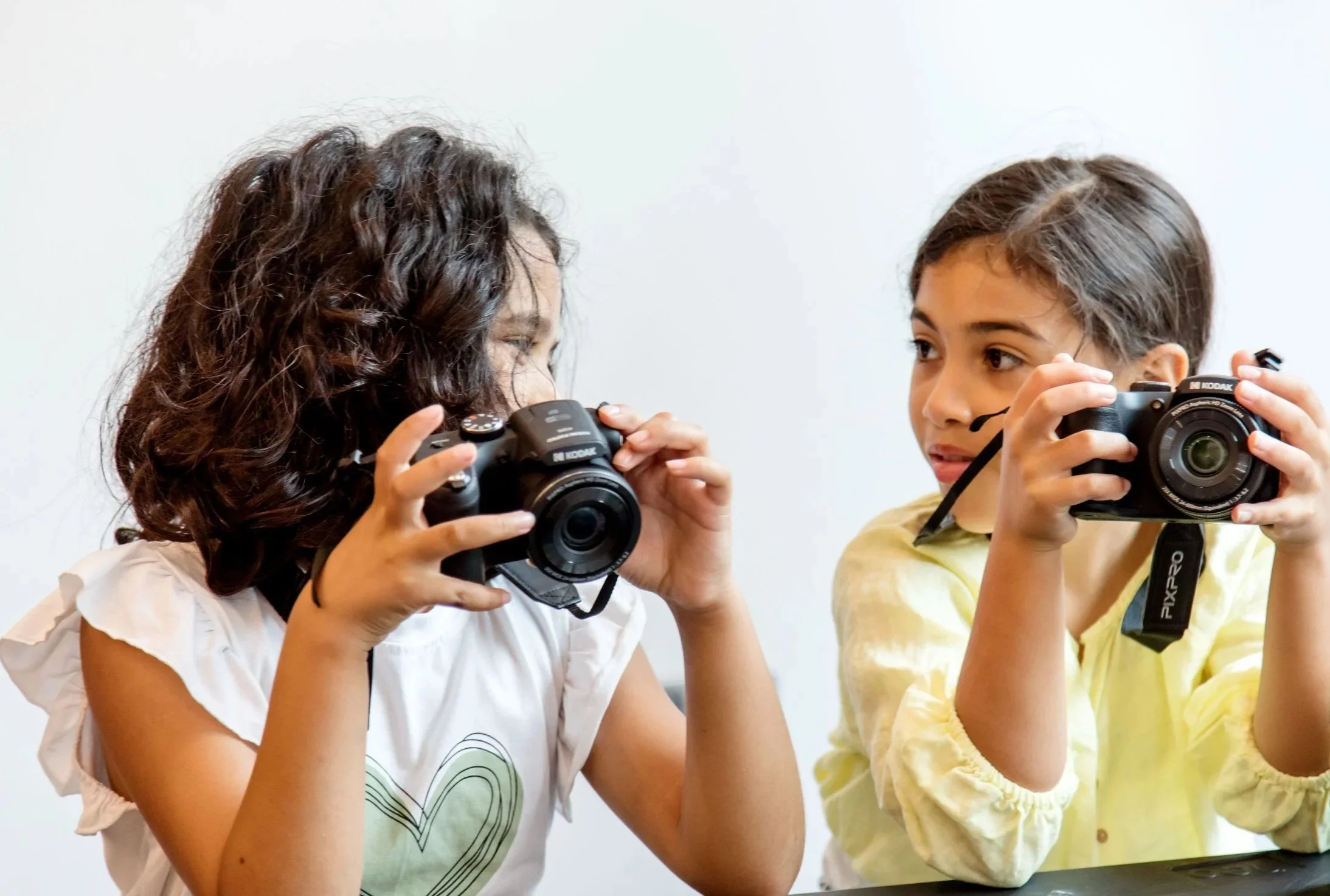 Two young girls are playing with cameras, one girl with curly hair wearing a white shirt and the other with straight hair in a yellow shirt, both holding cameras up to their faces.