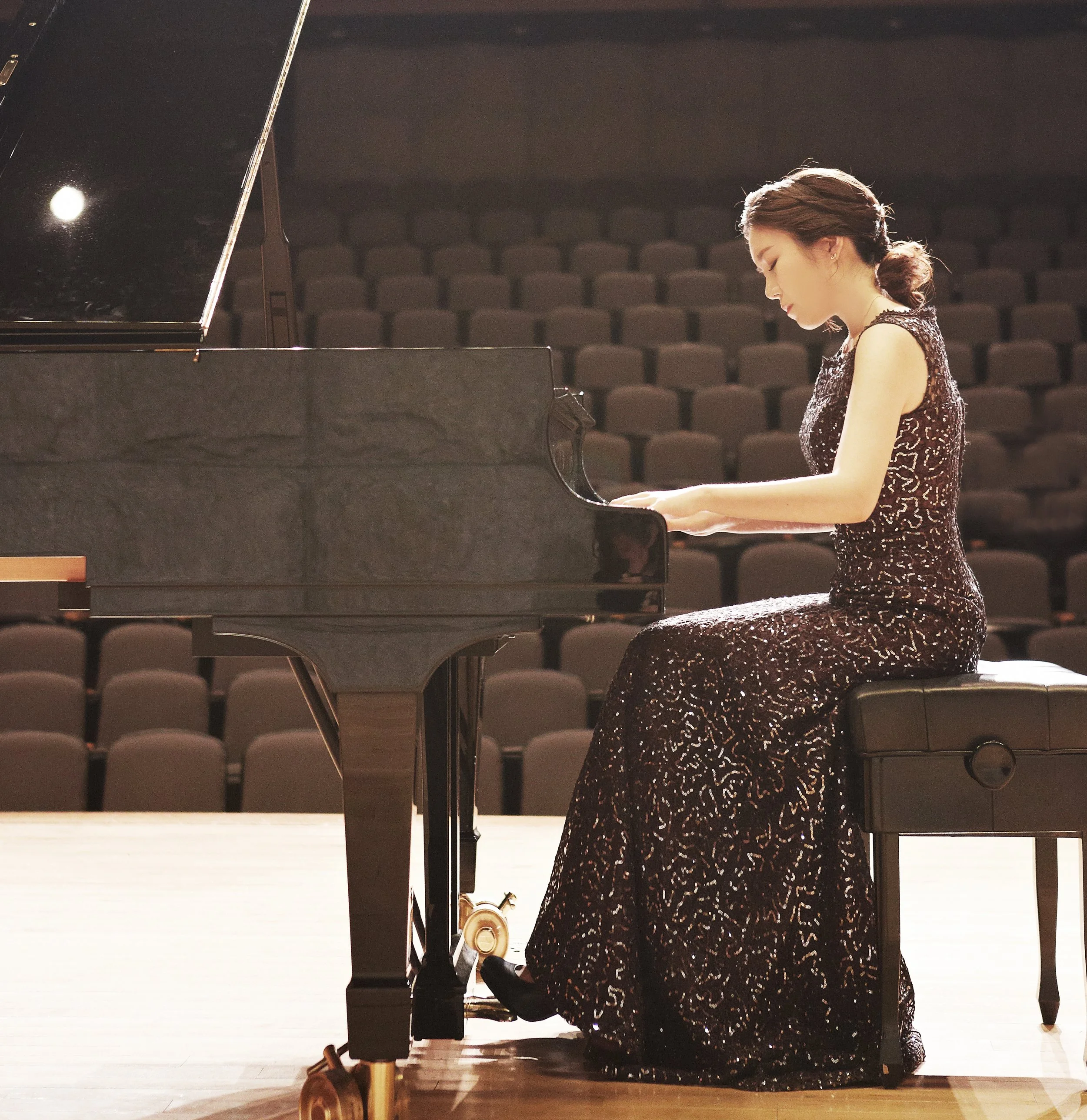 A woman in an elegant, dark, intricately patterned gown plays a grand piano on a stage in an empty concert hall.