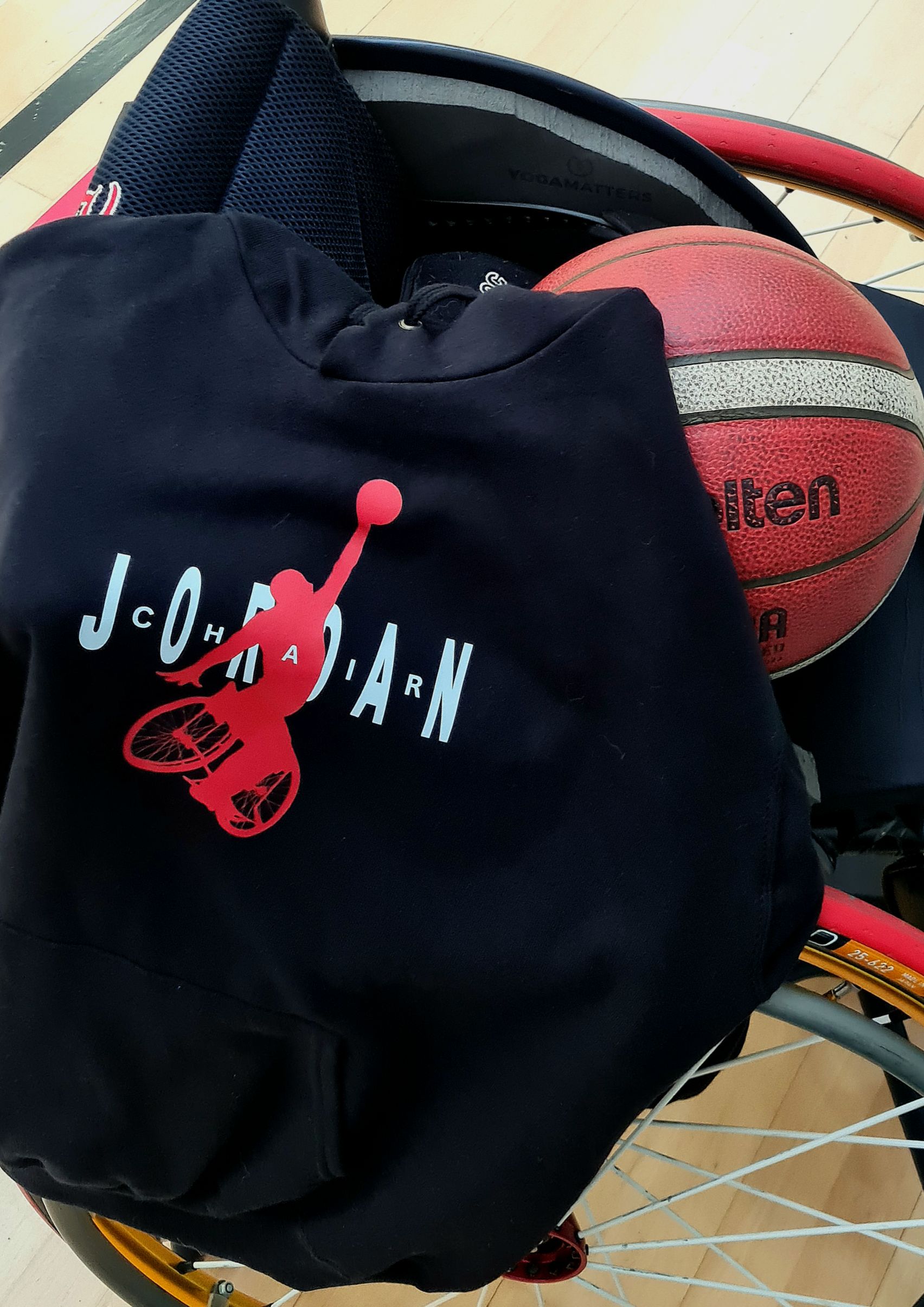 Close-up of a wheelchair with a basketball in the seat and a bag hanging on the side, on a wooden floor.