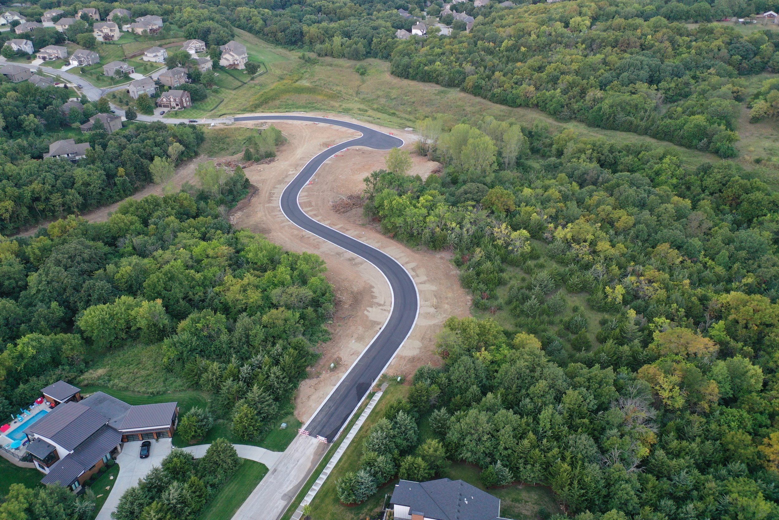 A winding road under construction in a verdant, wooded area with residential houses nearby.
