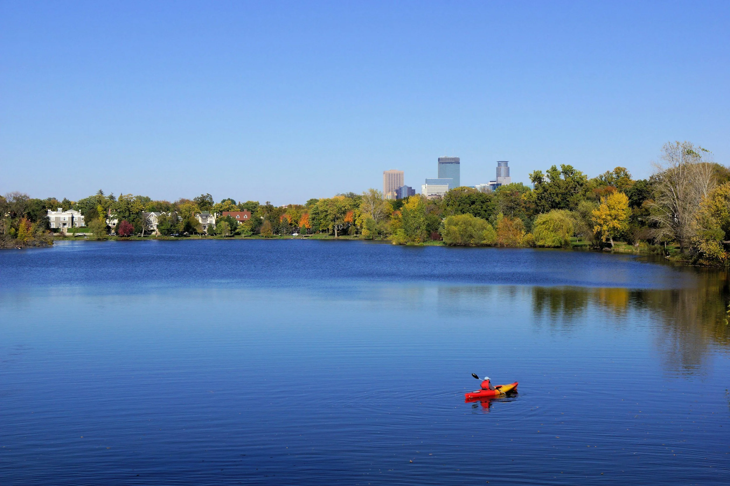 A red kayak with a person paddling on a calm lake with a city skyline in the background, surrounded by green and autumn-colored trees.