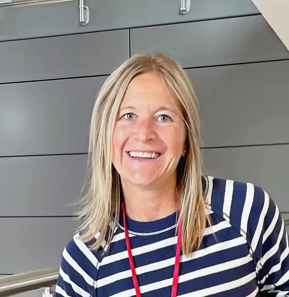 A woman with blonde hair, blue eyes, and a striped navy and white shirt smiling at the camera against a gray wall background.