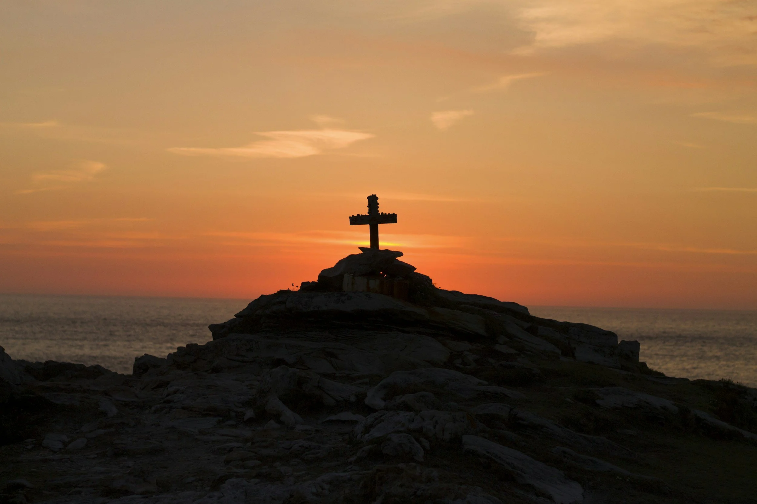 A cross-shaped monument on a rocky hill at sunset with the sky painted in warm orange and pink hues.