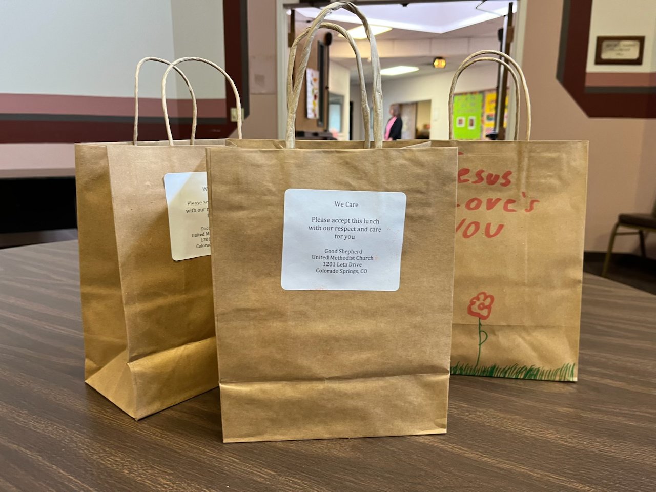 Three We Care brown paper lunch bags on a wooden table, with one featuring a white label with a prayer and church details, and another with a hand-drawn flower and message.
