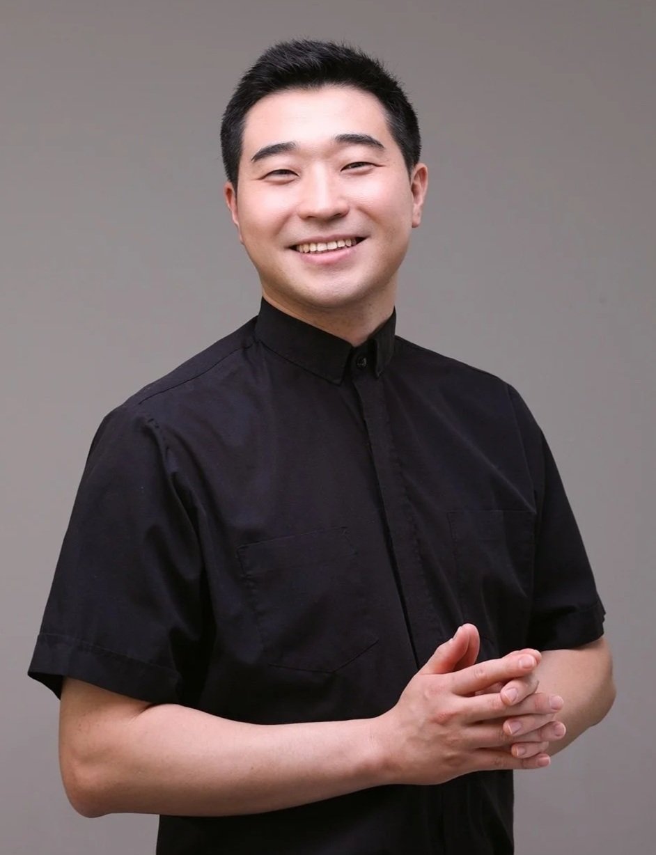 Pastor Peter Jisung Lee with short black hair and a bright smile, wearing a black short-sleeve button-up shirt, standing against a plain gray background.