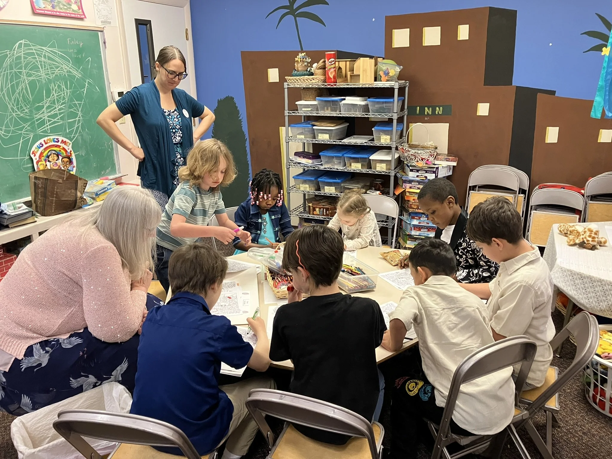 A group of children and an adult gather around a table, engaging in a craft activity in a classroom decorated with colorful murals, a green chalkboard, and organized shelves.