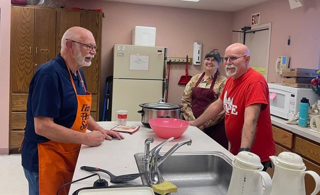 Good Shepherd Men's group hosting a pancake breakfast. Two men and one woman in the kitchen. The man on the left is wearing an orange apron, the woman in the middle is in a red apron, and the man on the right is in a red t-shirt.