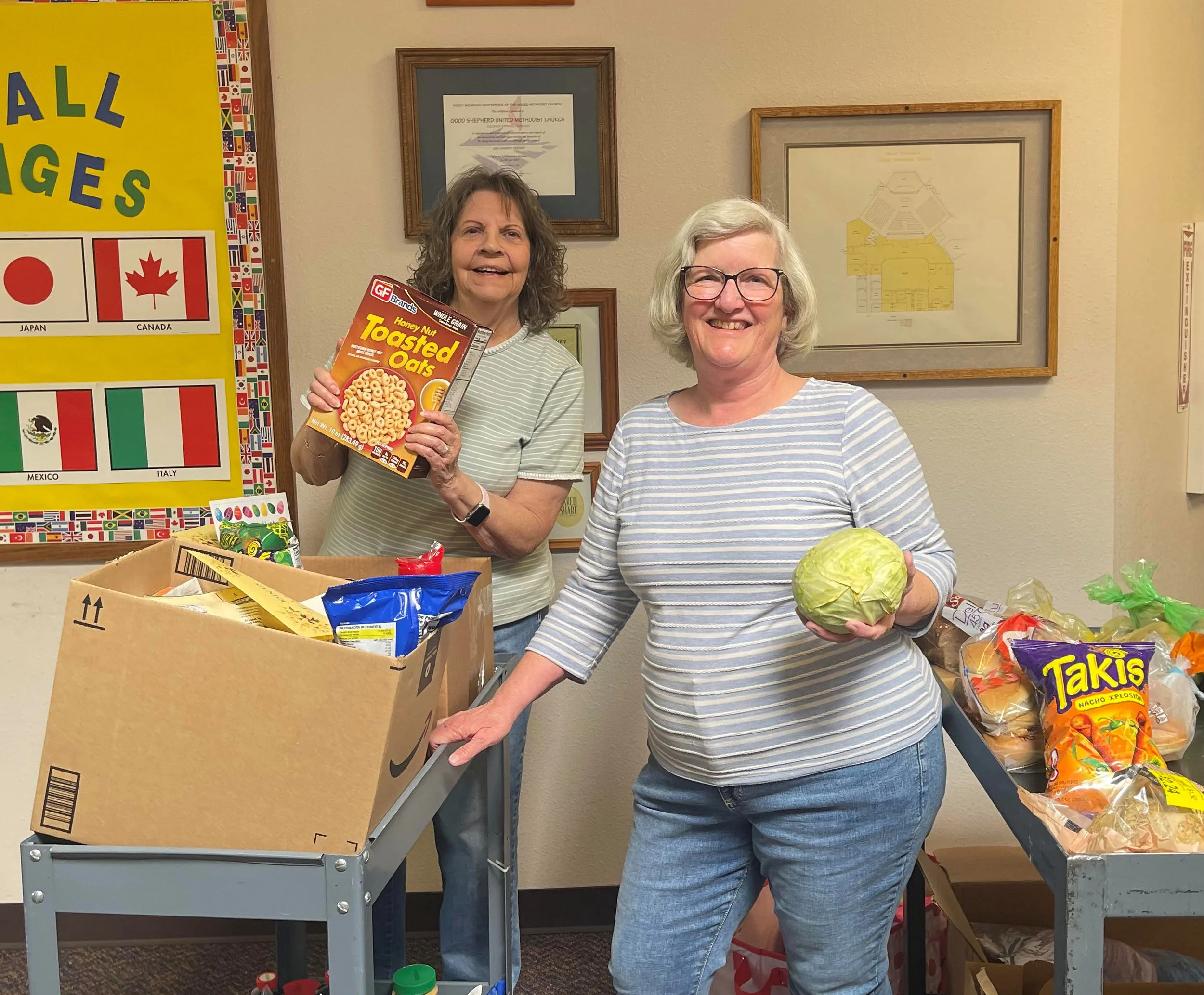 Two women working the Emergency Food Pantry are shelf-stocking and handing out food. One woman is holding a box of Honey Nut Toasted Oats cereal, and the other is holding a head of cabbage.