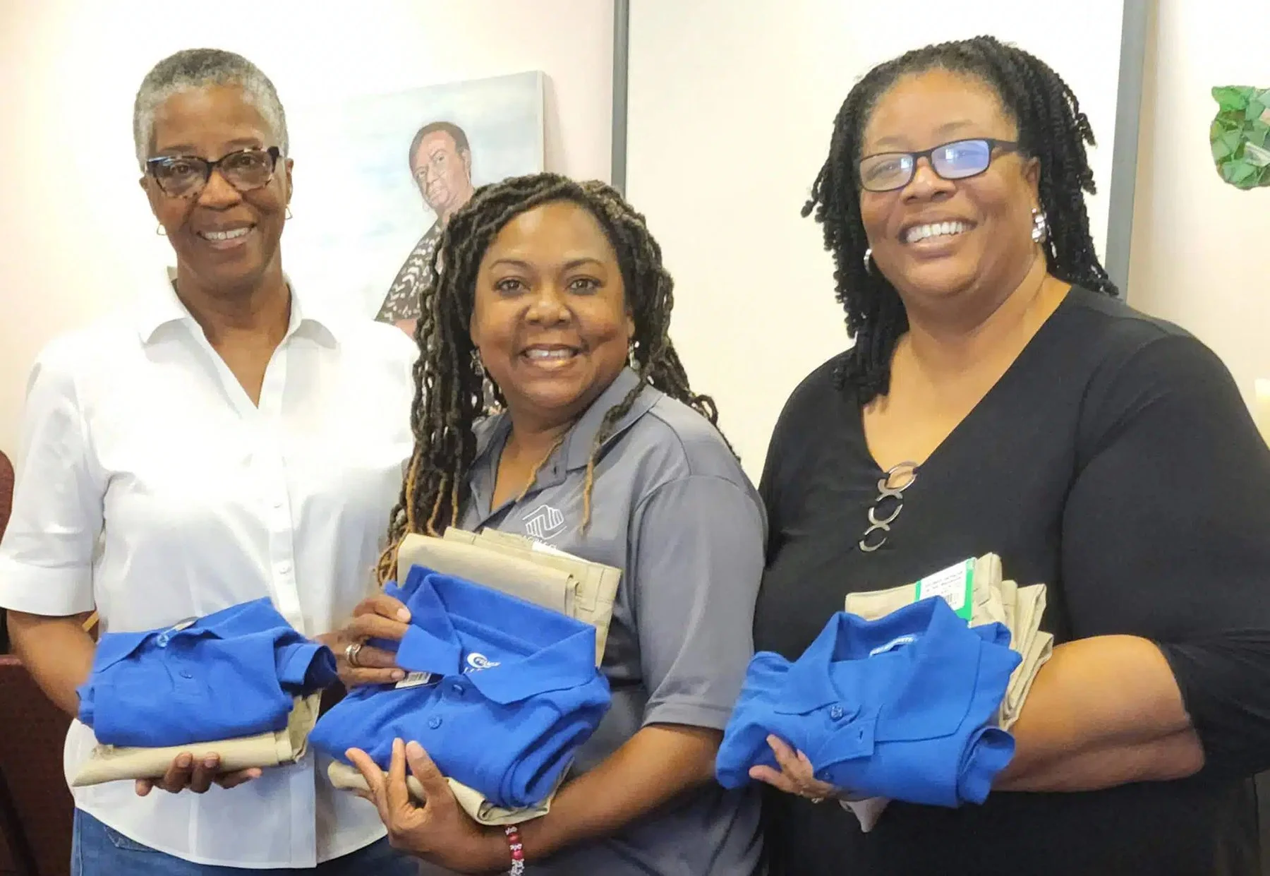 Three women holding blue tote bags and papers, standing indoors with a painting of a man in the background. All are smiling and dressed in casual business attire.