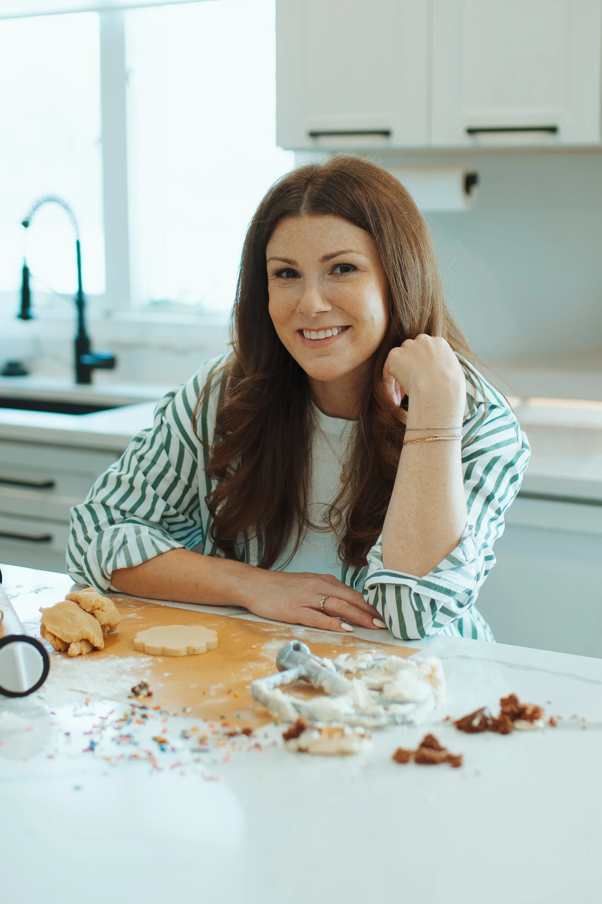 A woman with long brown hair smiling at the camera while sitting at a kitchen counter surrounded by baking supplies and cookies.