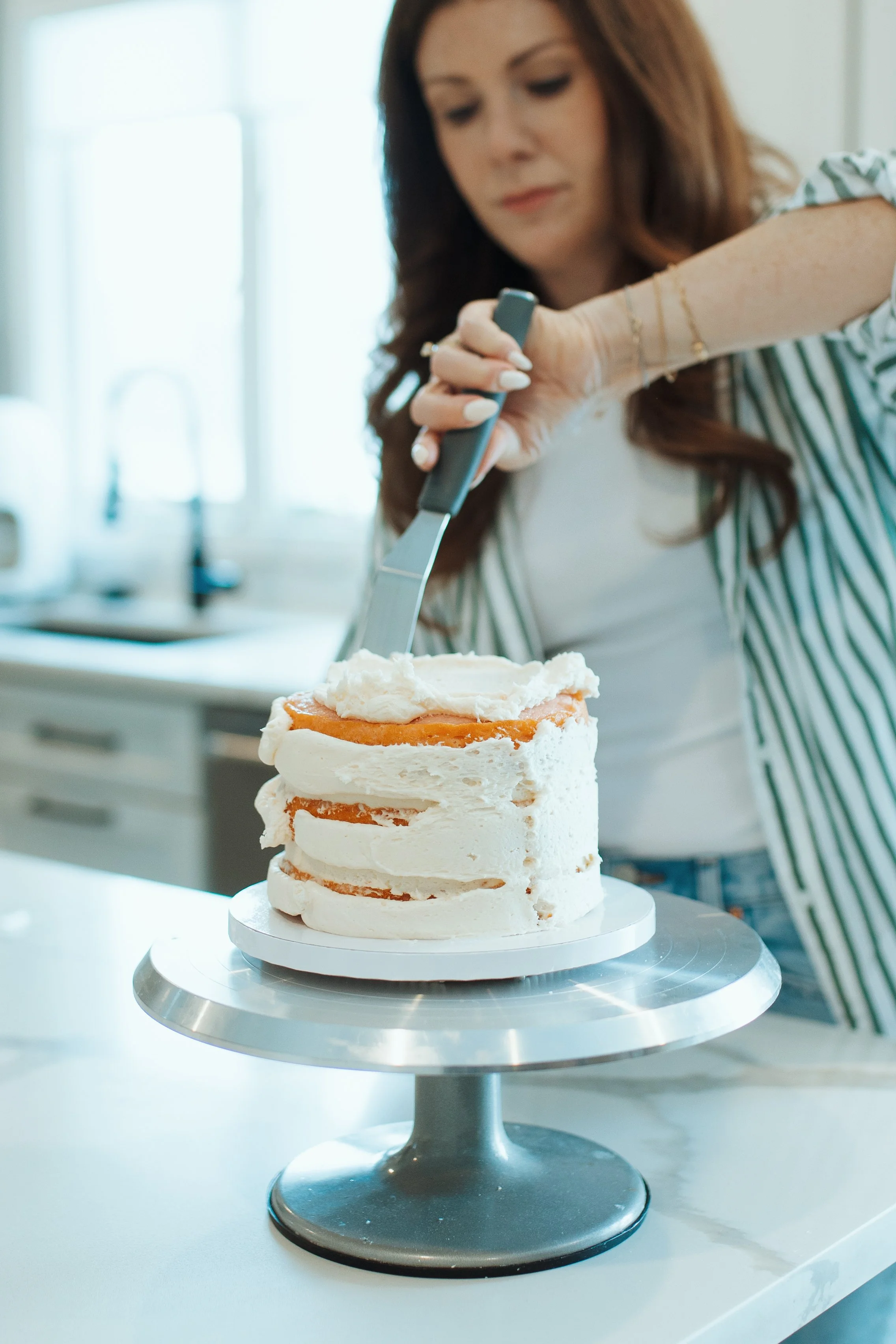 A woman decorating a layered cake with white frosting in a kitchen with natural light.