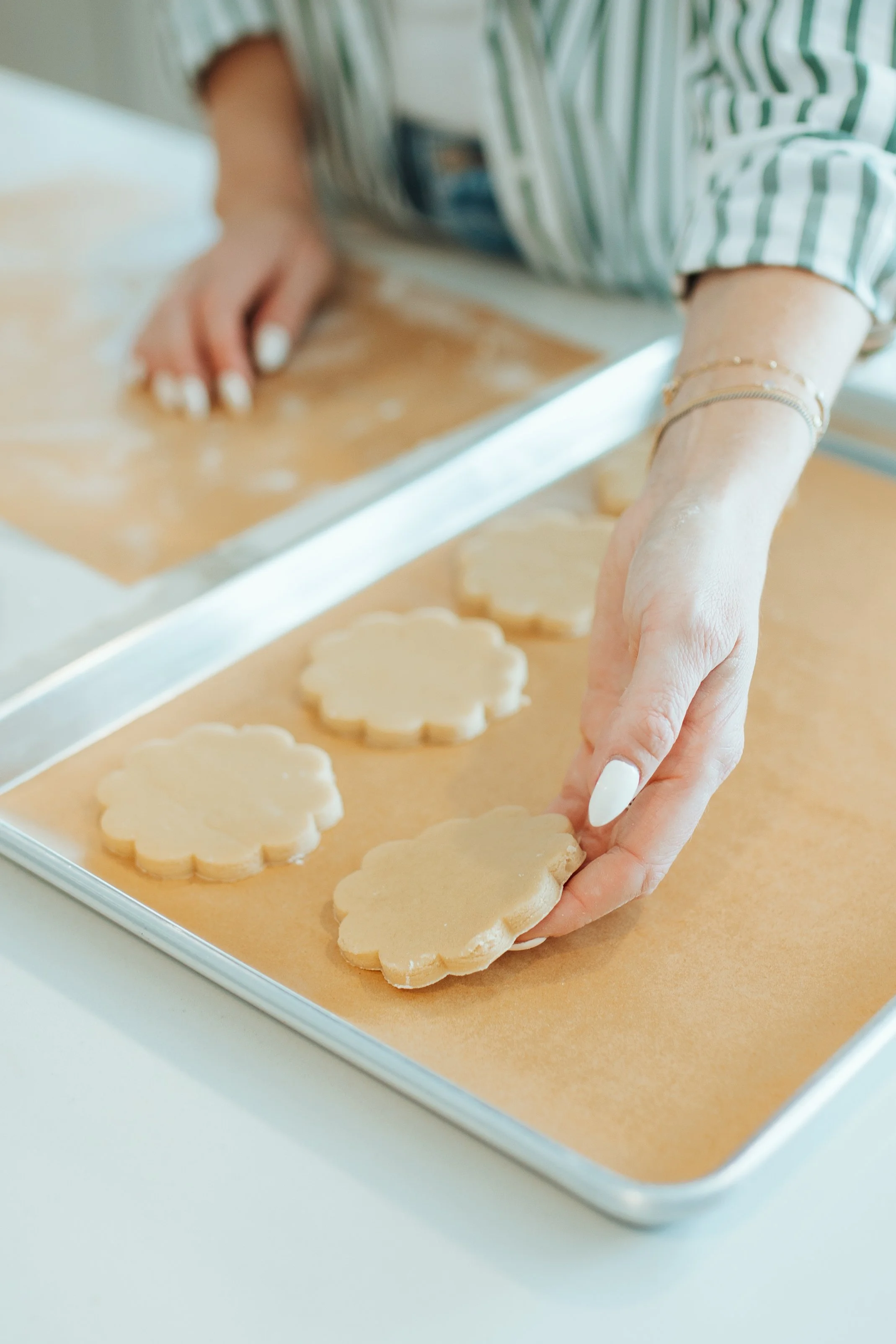 Person's hand placing flower-shaped cookie cutouts on a baking sheet with parchment paper, with more cookies in the background.