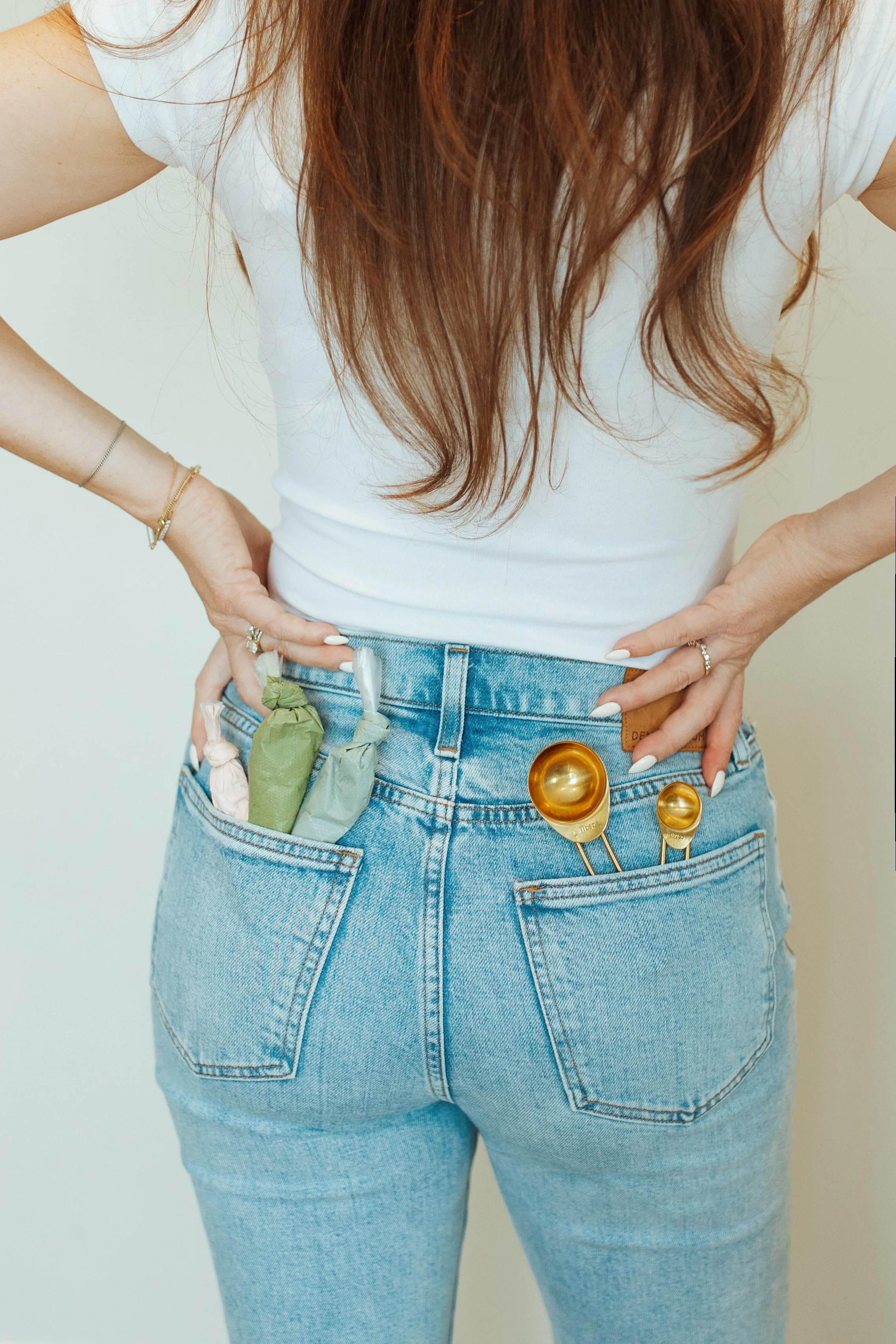 Back view of a woman in a white shirt and light blue jeans with a pocket filled with salon tools, including brushes and color tubes.