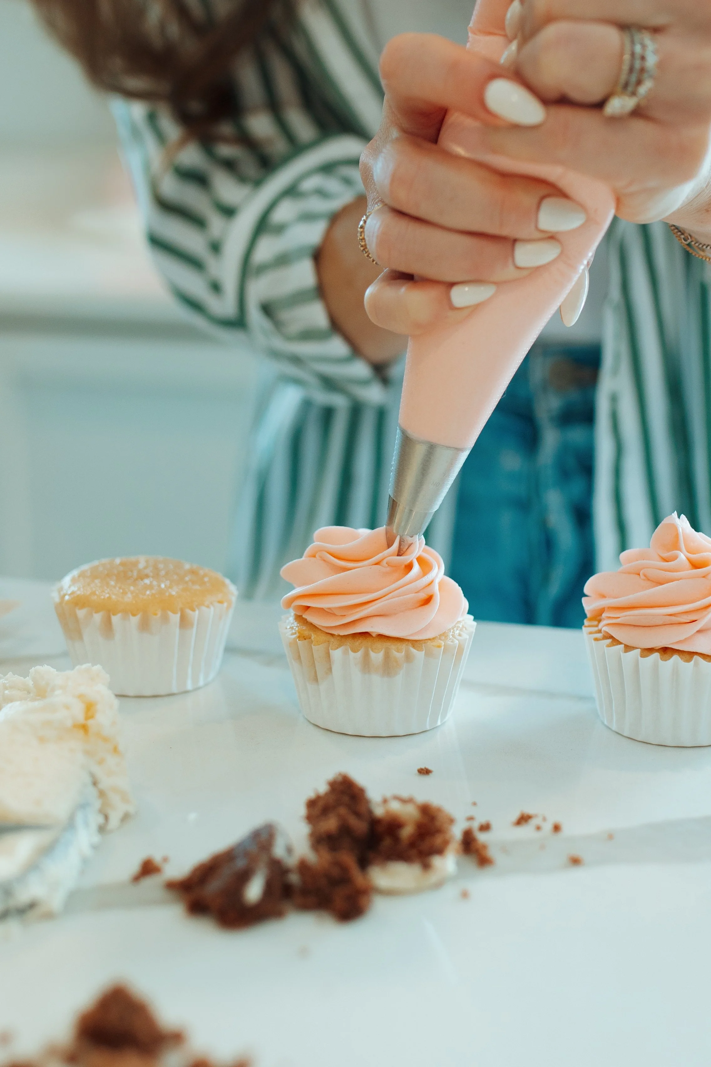 A person decorates cupcakes with pink frosting using a piping bag. Several cupcakes and cake crumbs are on the table.