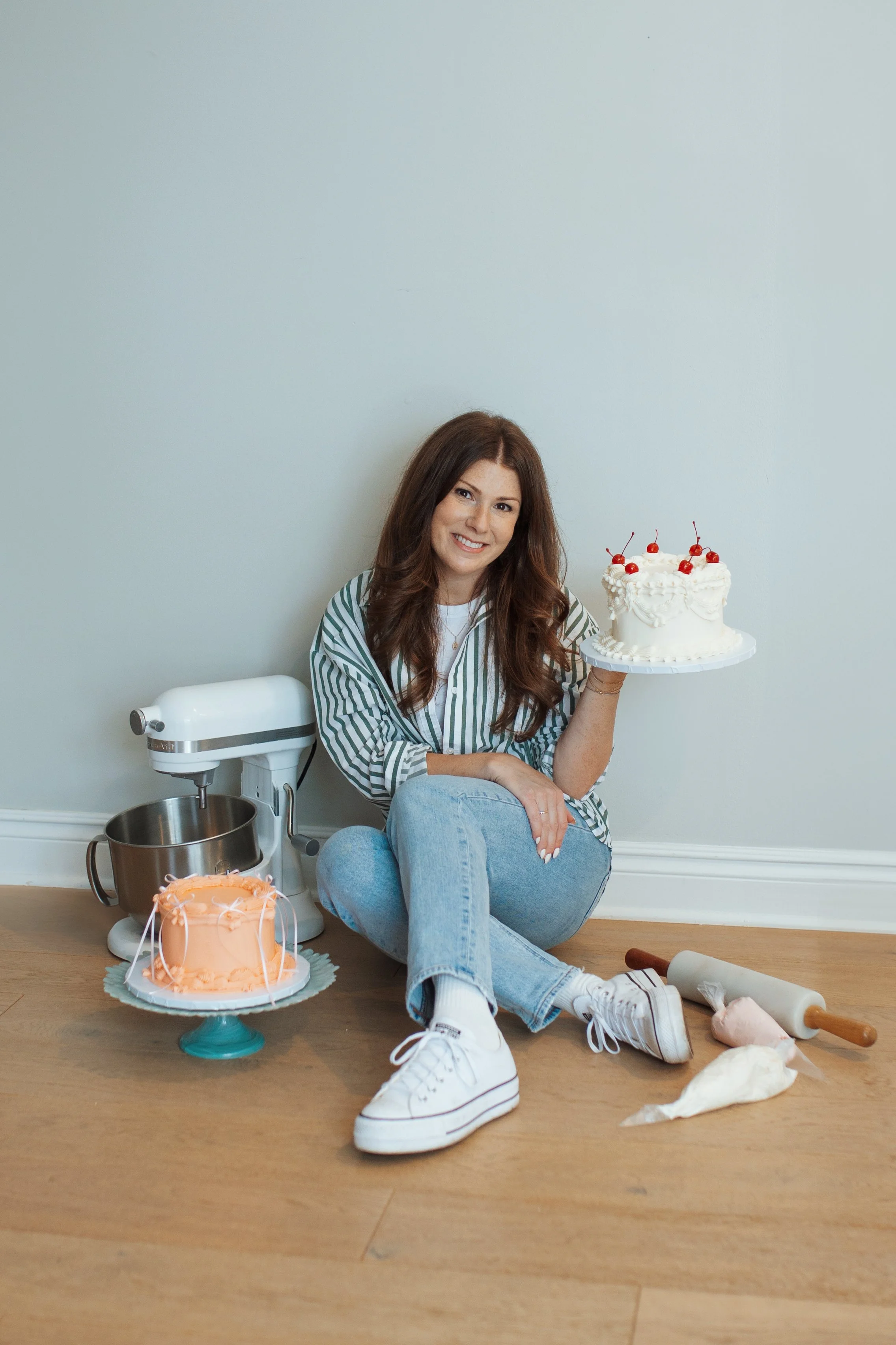 A woman sitting on the floor with a smile, holding a white cake topped with cherries. There are two other cakes, a stand mixer, a rolling pin, and baking supplies around her. The setting is a simple room with a light-colored wall and wooden floor.