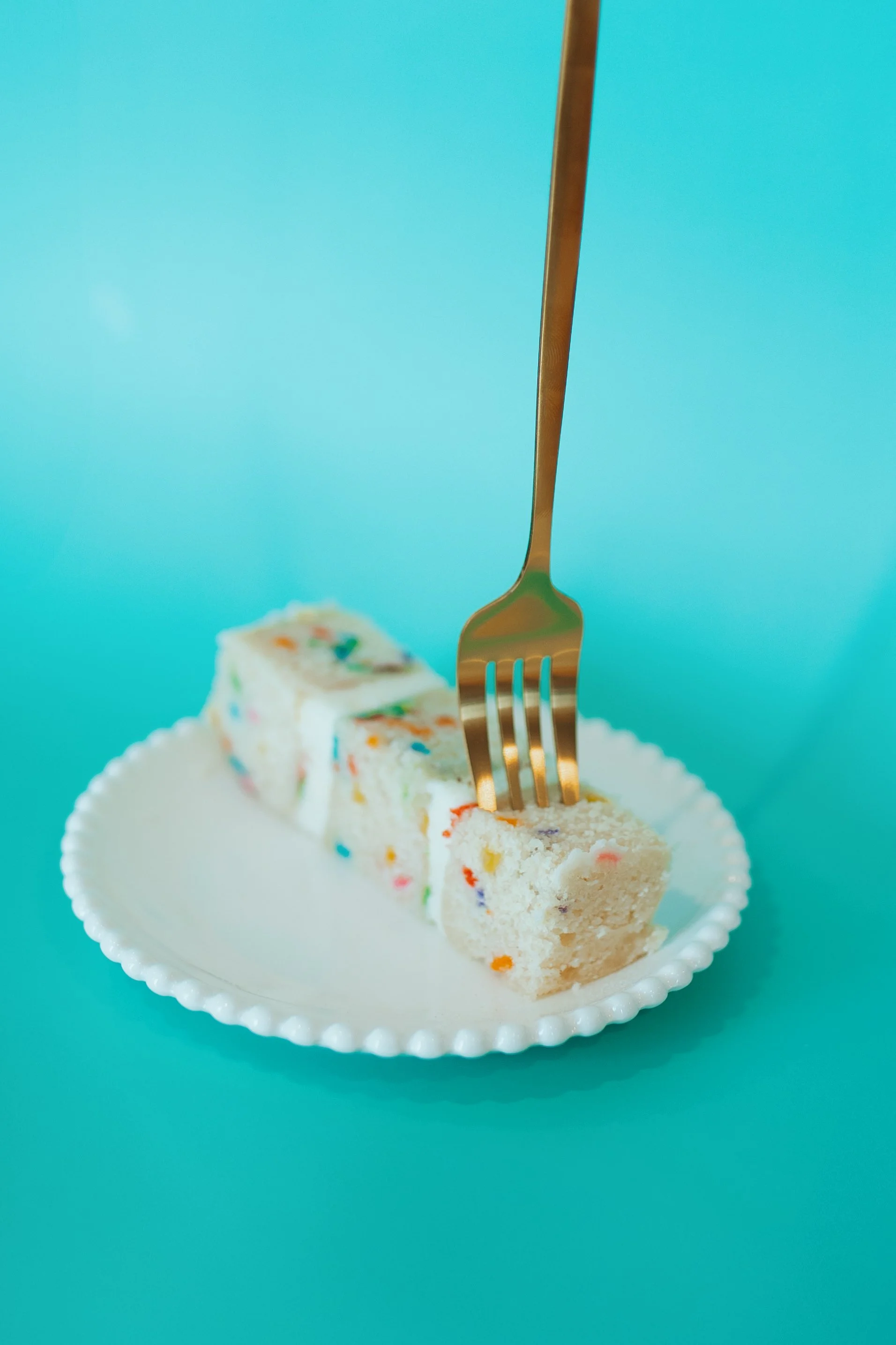 Slices of colorful birthday cake on a white plate with a fork, set against a turquoise background.
