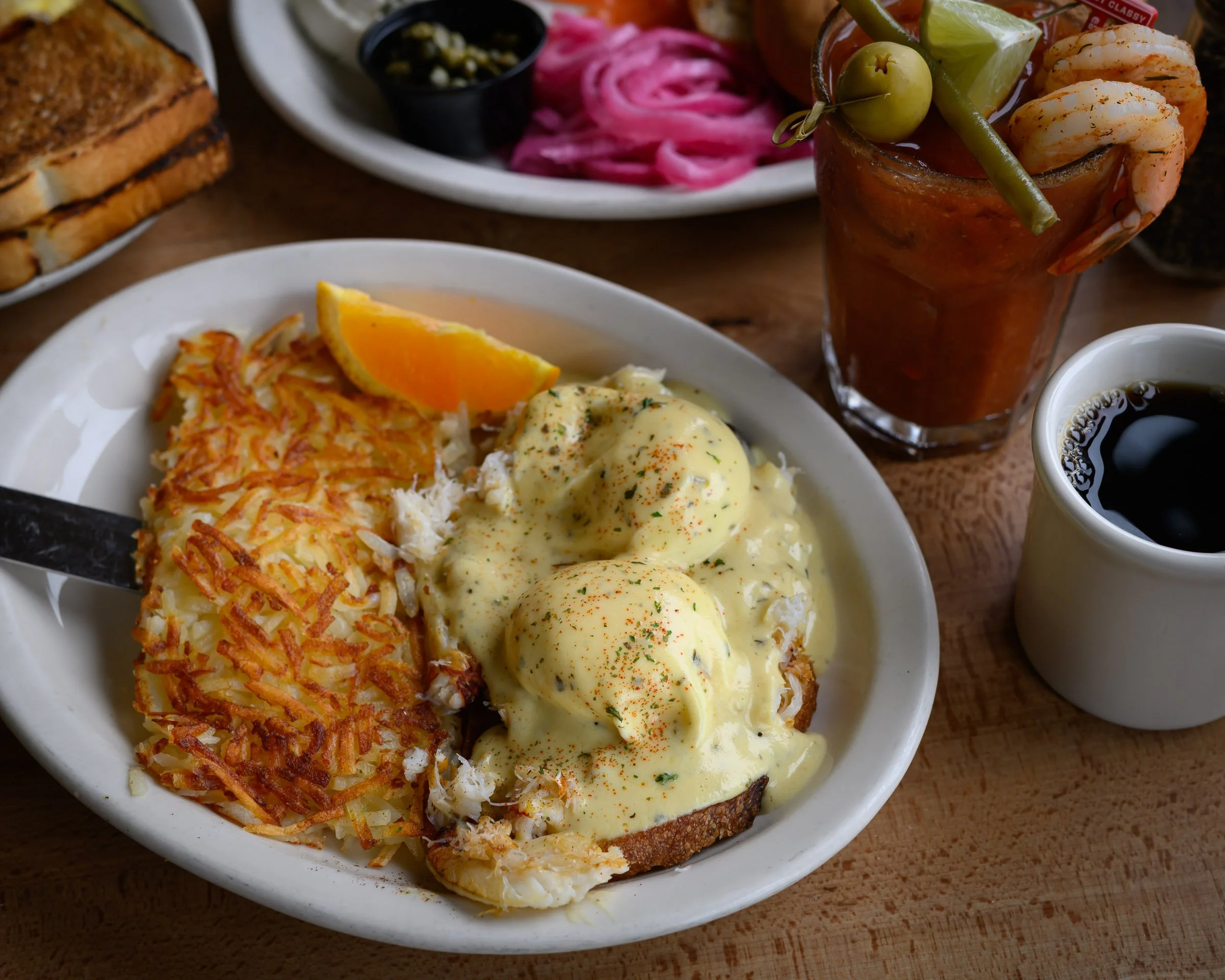 Plate with eggs Benedict topped with hollandaise sauce, served with hash browns and an orange slice. In the background, there is a Bloody Mary garnished with shrimp, olives, a lime wedge, and green beans; a small cup of black coffee; pink pickled oni