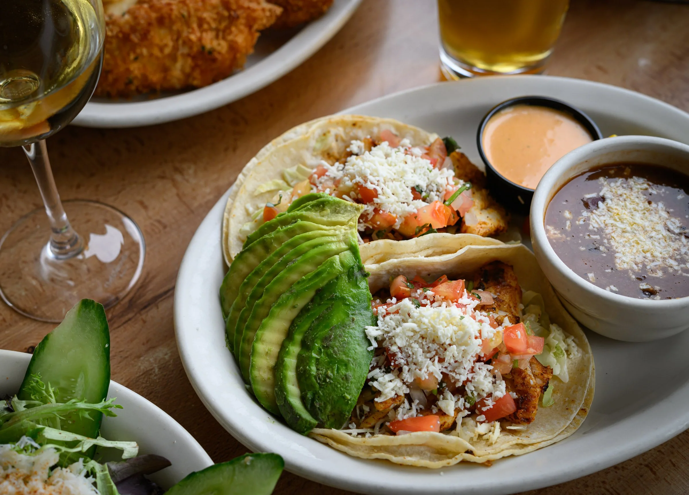 Close-up of a plate with two soft shell tacos filled with chopped tomatoes, shredded cheese, and lettuce, garnished with sliced avocado. In the background, there are bowls of black beans and an orange sauce, with a glass of beer and a plate with frie